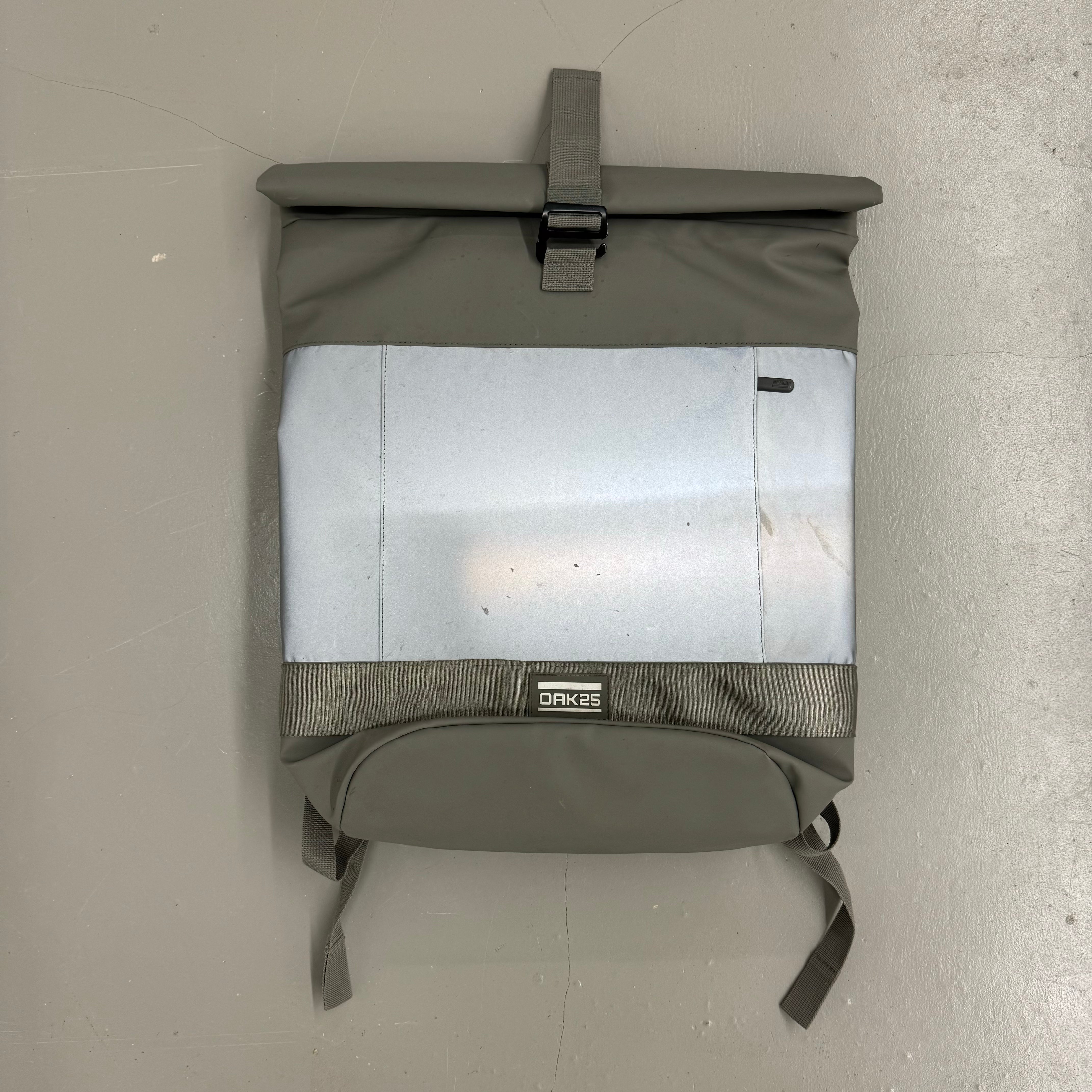 Ein grauer Rucksack mit einer hellen Fronttasche und einem Rollverschluss liegt auf einem grauen Boden.