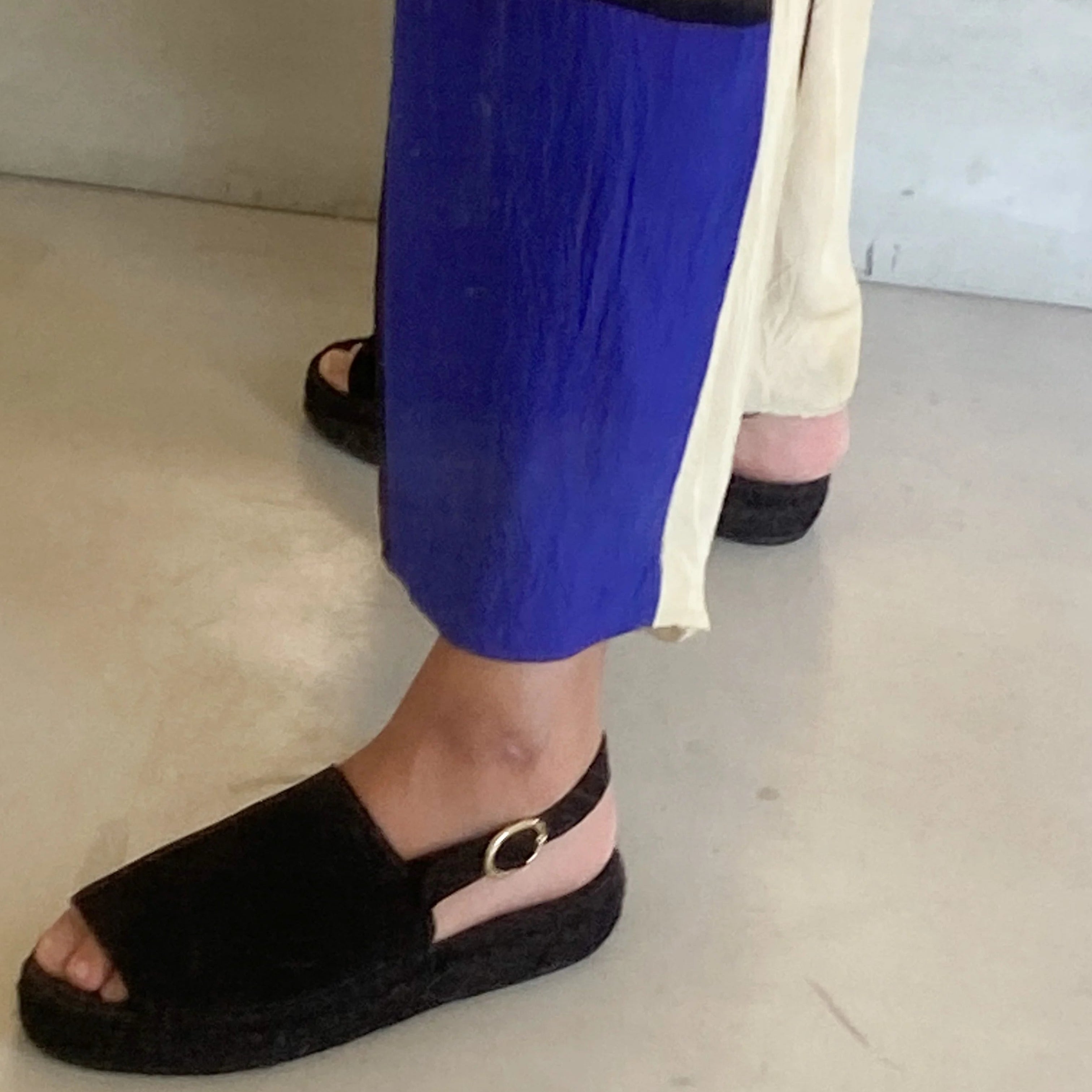 Eine Person, die eine cremefarbene und blaue Hose trägt, steht auf einem hellen Boden und zeigt schwarze Plateausandalen mit offenen Zehen und Knöchelriemen.