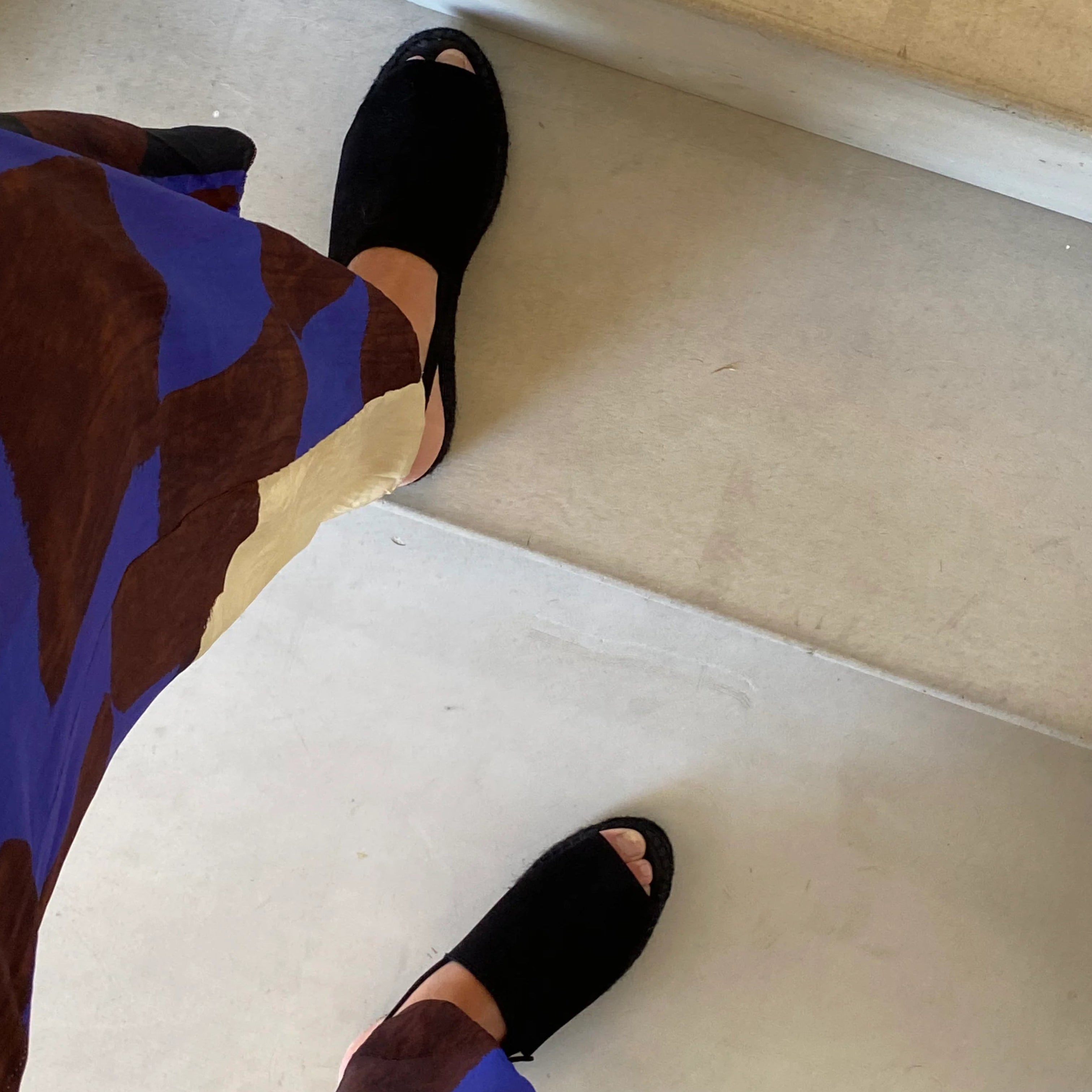 Eine Person, die schwarze offene Schuhe und eine weit geschnittene Hose mit blauen, braunen und beigen Mustern trägt, betritt eine helle Treppe.