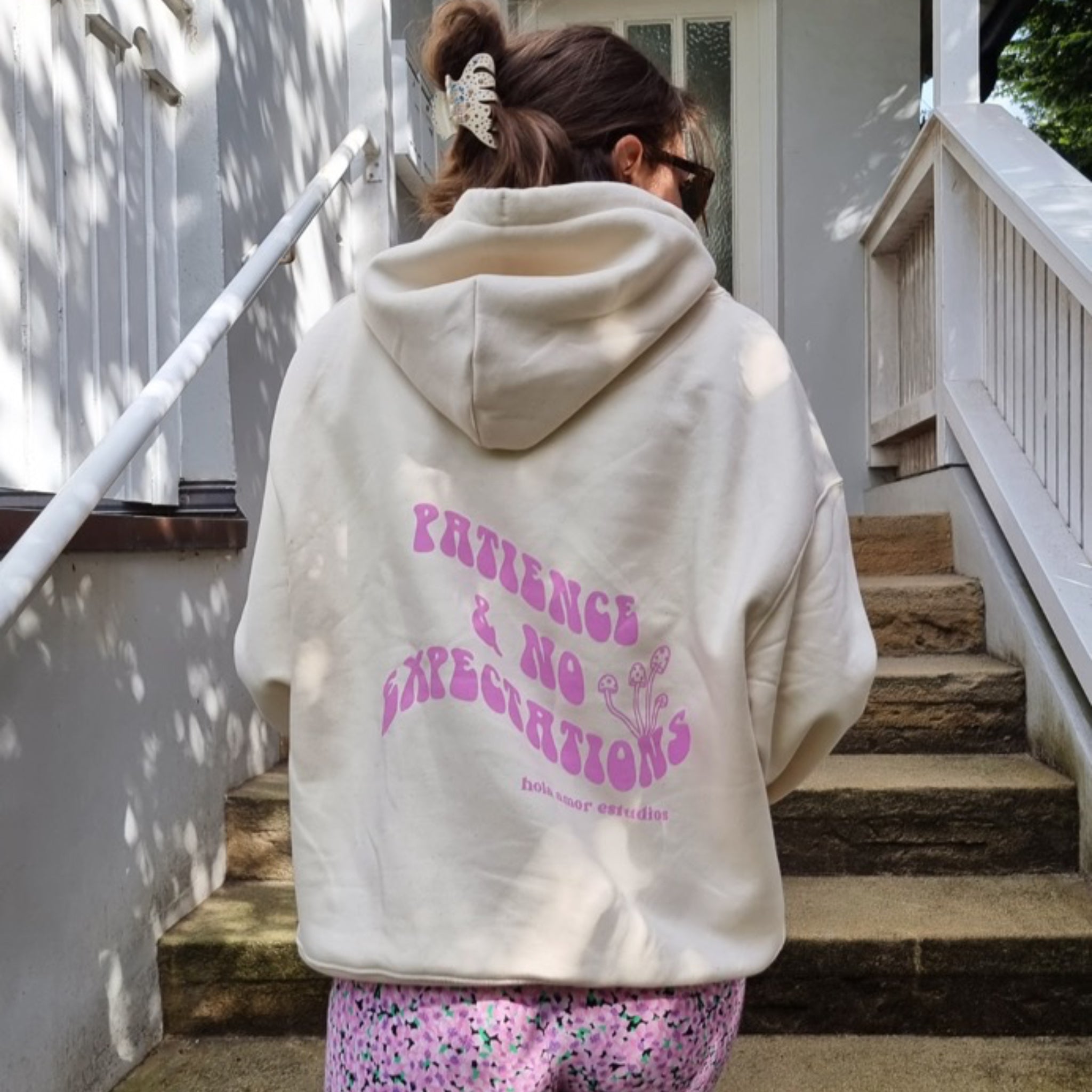 Frau mit Sonnenbrille auf Treppe, trägt weißen Hoodie mit Aufschrift "Patience & No Expectations" in rosa Buchstaben.