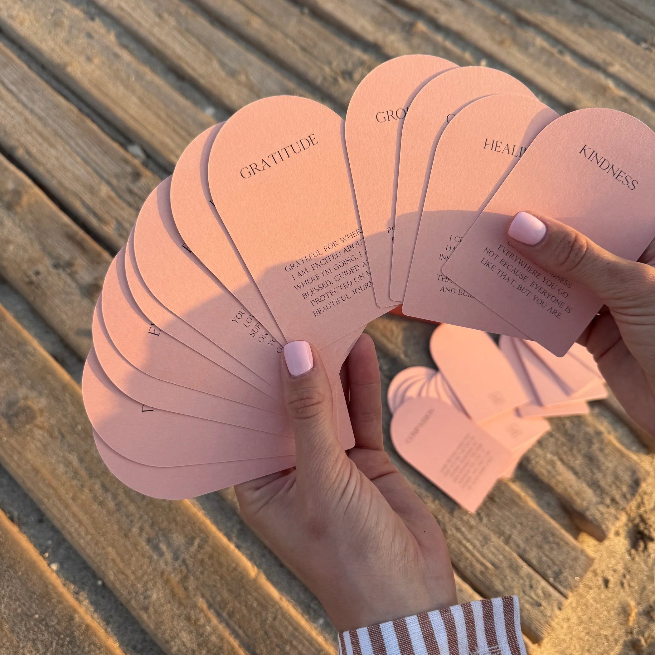 Eine Person mit blassrosa Nägeln hält einen aufgefächerten Stapel rosafarbener Karten, auf denen Worte wie GRATITUDE und KINDNESS stehen. Die Person steht auf einer Holzfläche im Freien.