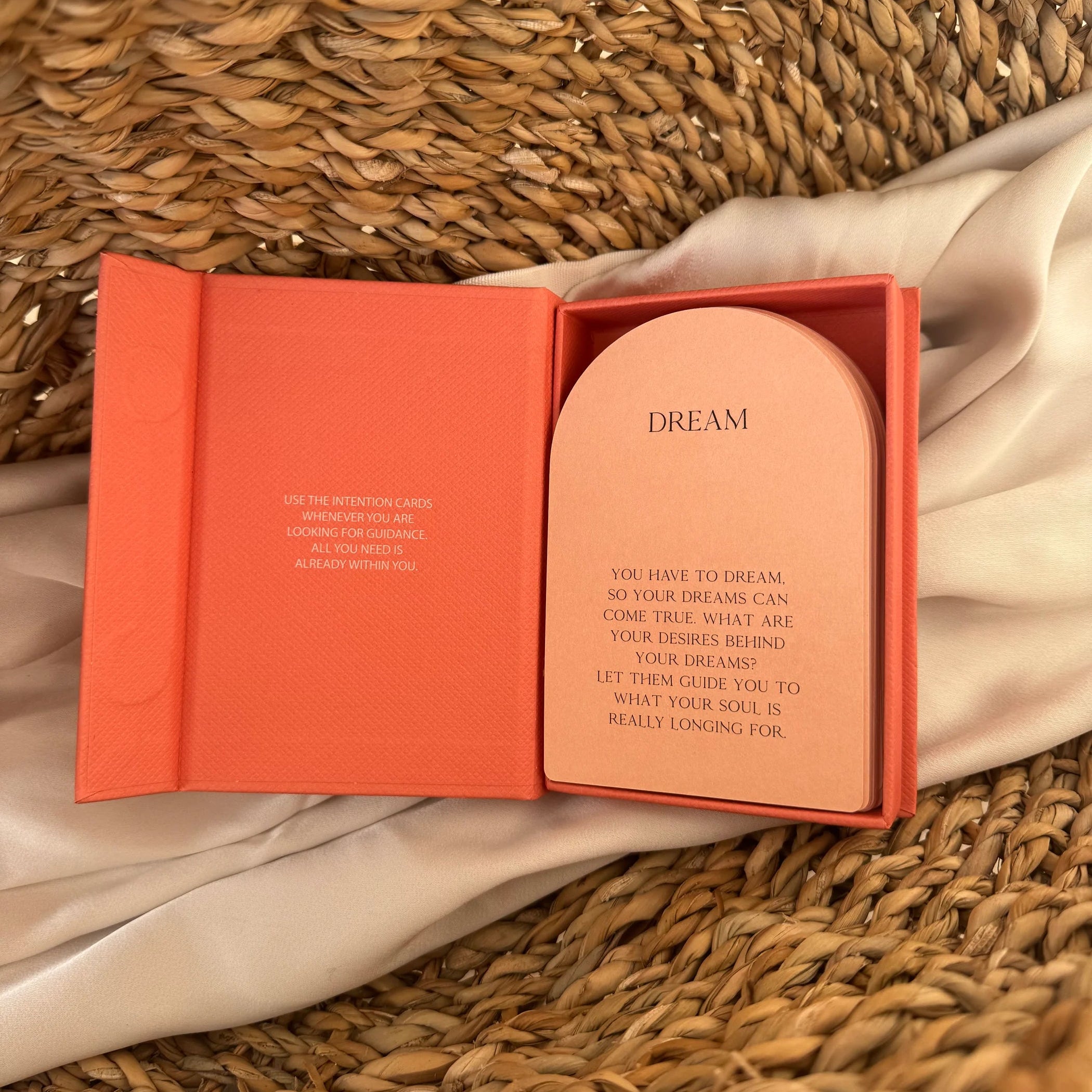 Eine orangefarbene Schachtel mit Absichtskarten liegt geöffnet auf geflochtenem Stroh mit einem Satintuch und enthüllt eine Karte mit der Aufschrift DREAM und einer darauf gedruckten Botschaft über Träume und Wünsche.