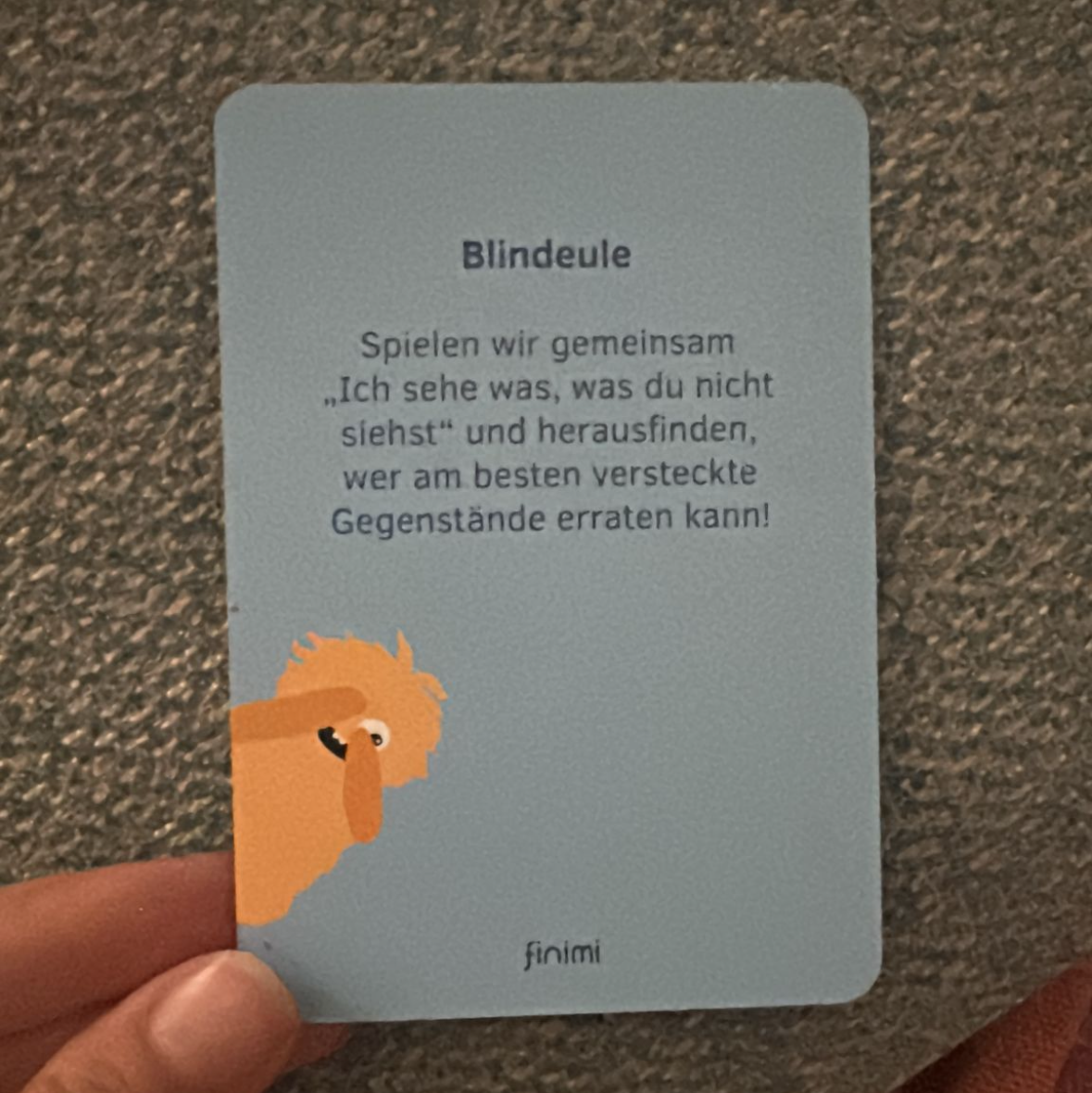 Eine Hand hält eine blaue Karte mit deutschem Text und einer Cartoon-Eulenillustration in der unteren linken Ecke. Die Karte erklärt ein Spiel namens Blindeule, bei dem es darum geht, versteckte Gegenstände zu finden.