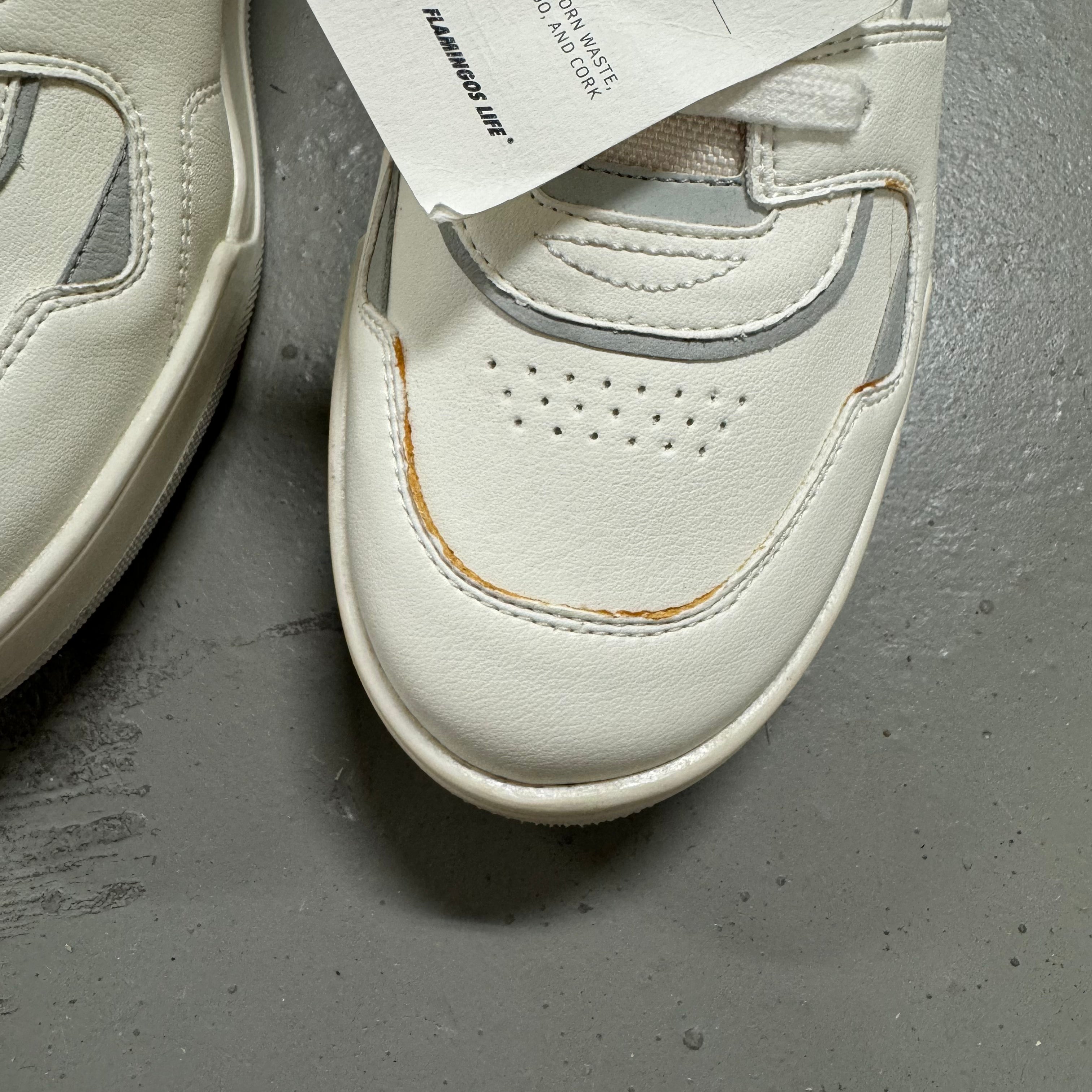 Weiße Sneaker mit grauen Akzenten auf einem grauen Boden; ein Etikett von "Flamingos Life" ist angehängt.