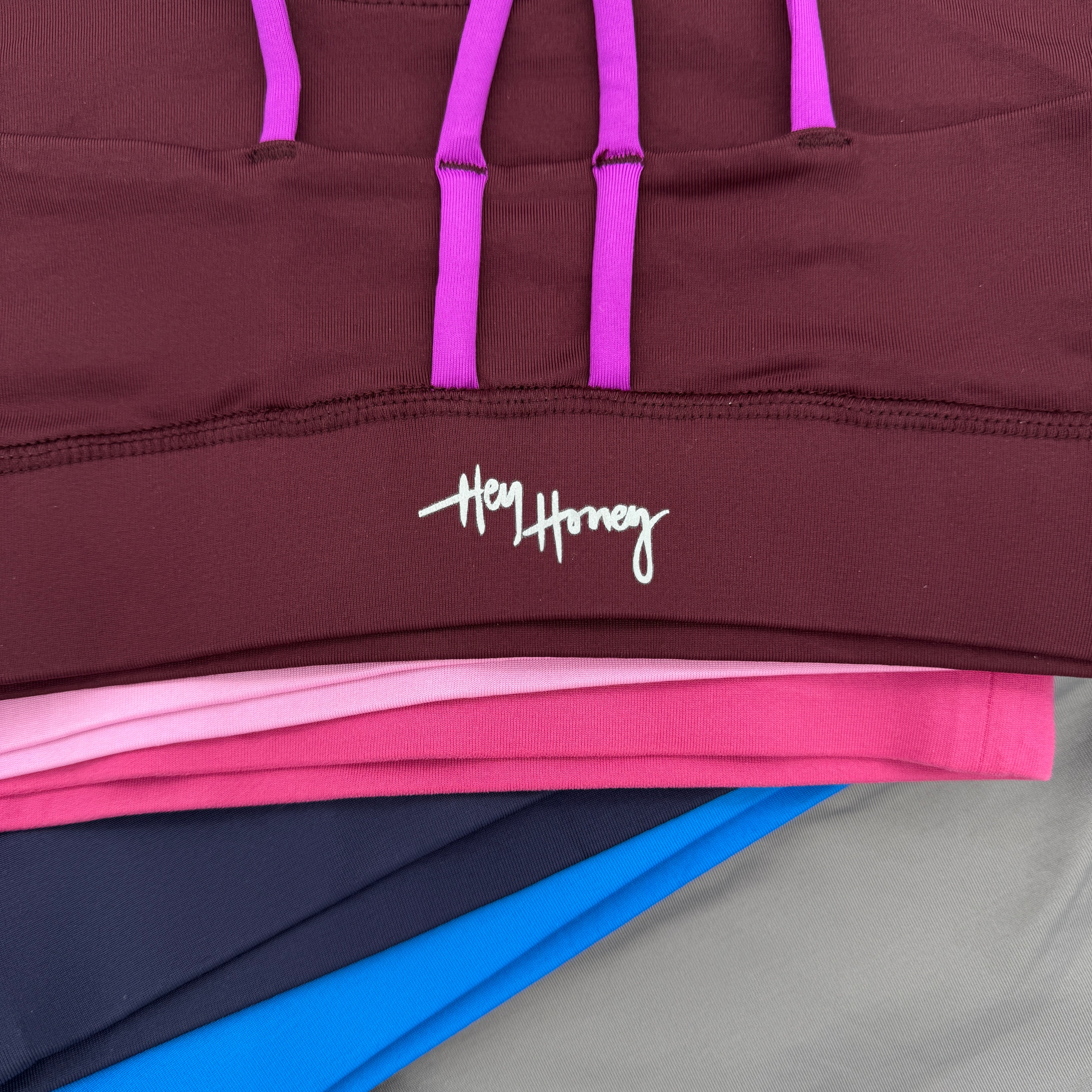 Eine Nahaufnahme von gefalteter Sportkleidung in verschiedenen Farben, darunter Burgunderrot, Rosa, Marineblau, Blau und Grau. Auf dem weinroten Teil oben ist das Logo Hey Honey in Weiß aufgedruckt.
