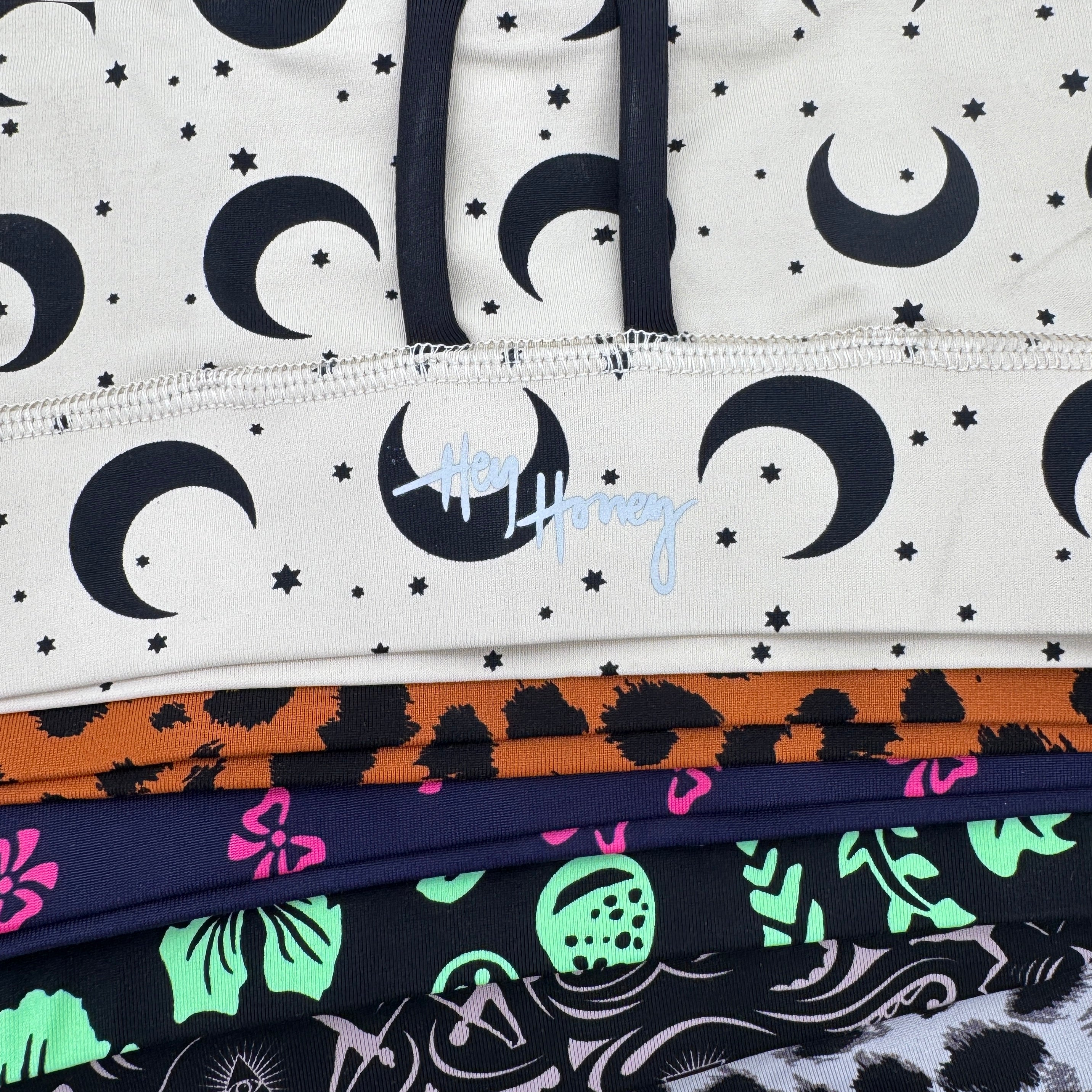 Ein Stapel gefalteter Leggings mit verschiedenen Mustern, darunter cremefarben mit schwarzen Monden und Sternen, orange und schwarz mit Leopardenmuster, marineblau mit Blumen, neongrün und rosa tropisch und schwarz mit weißen Skeletten.