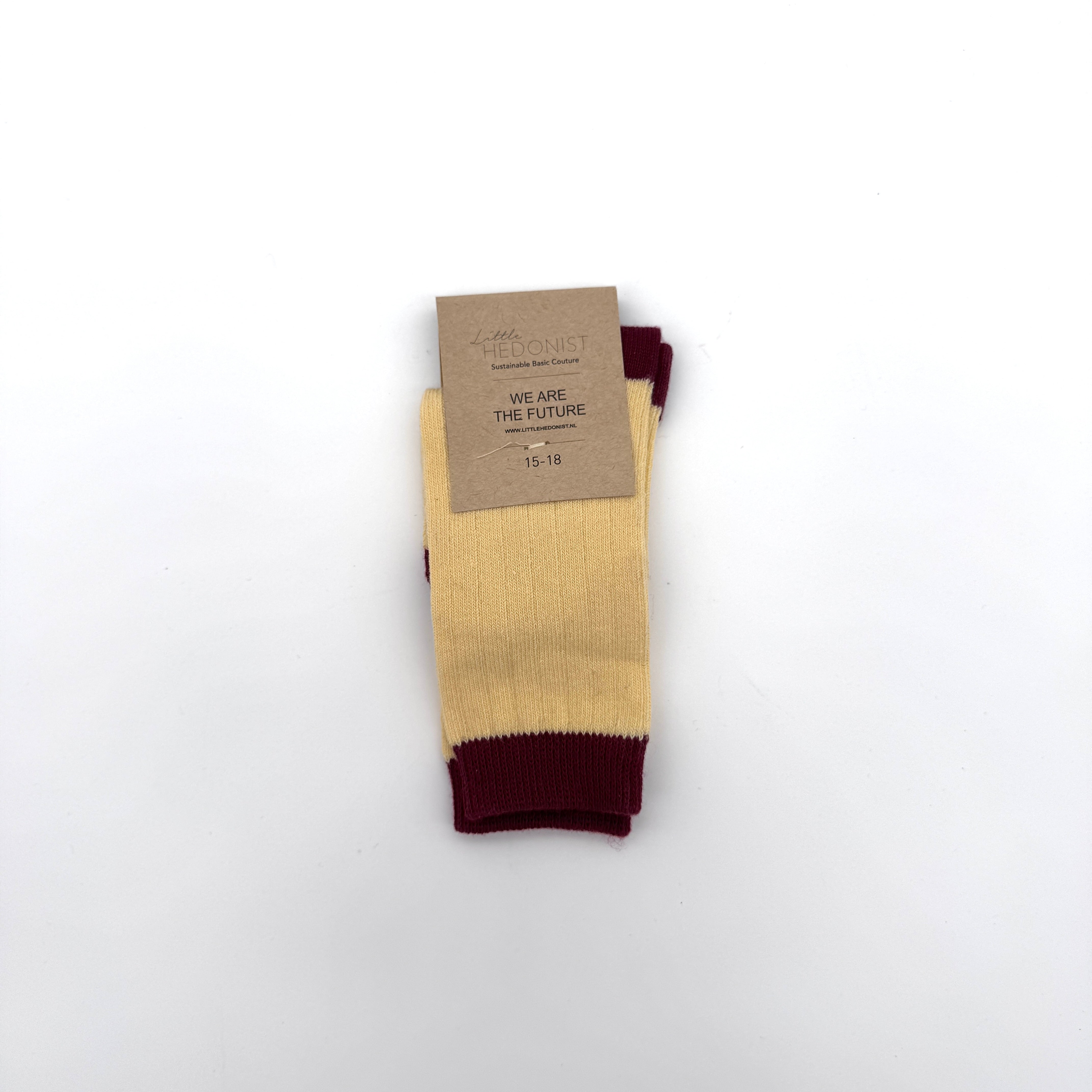 Ein Paar gerippte gelbe Socken mit roten Bündchen, Zehen und Fersen, ordentlich gefaltet mit einem braunen Kartonetikett mit der Aufschrift Little Hedonist - WE ARE THE FUTURE - 15-18 auf weißem Hintergrund.