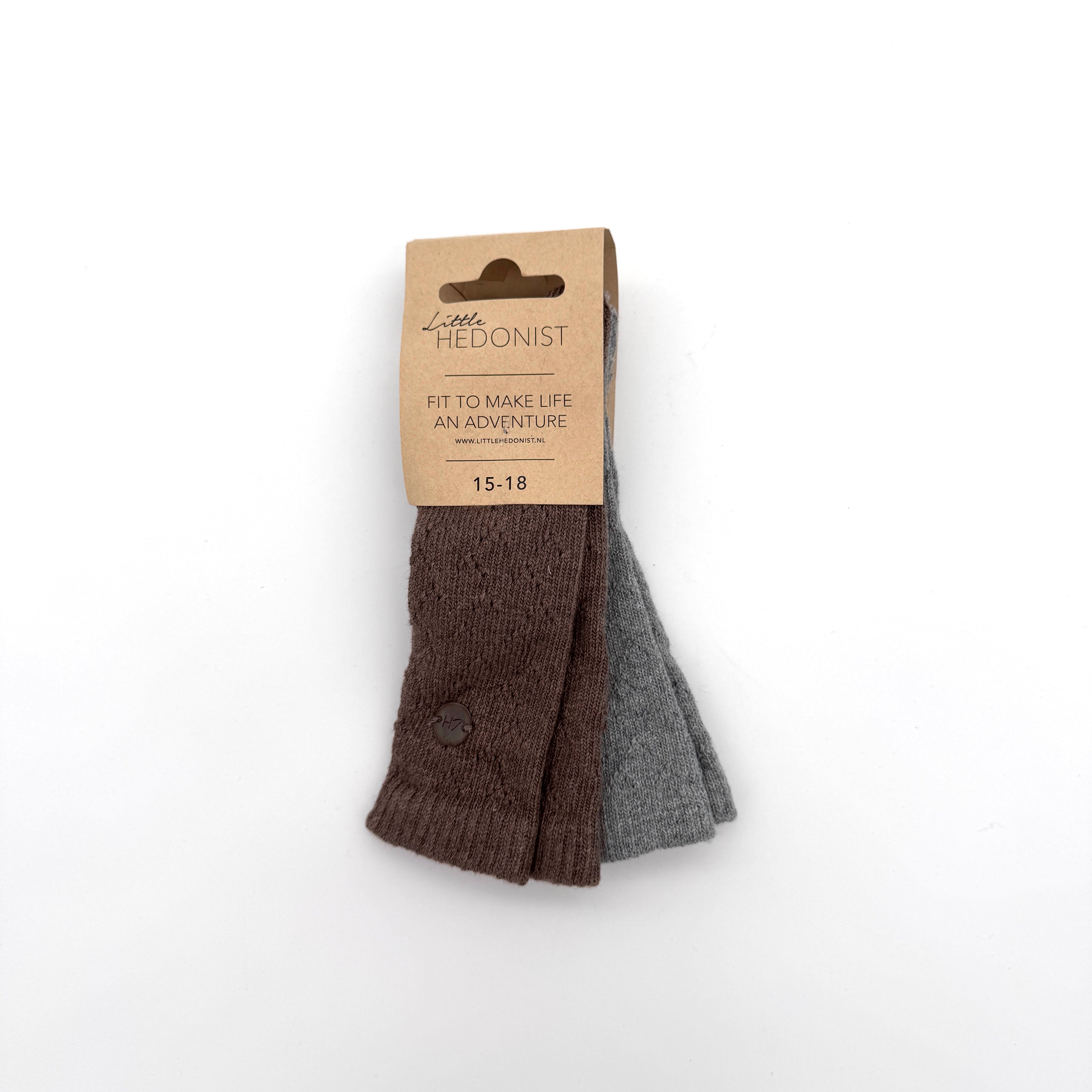 Ein Paar Kindersocken, eine braune und eine graue, mit einem Kartonetikett mit der Aufschrift Little HEDONIST, FIT TO MAKE LIFE AN ADVENTURE, 15-18, auf weißem Hintergrund.