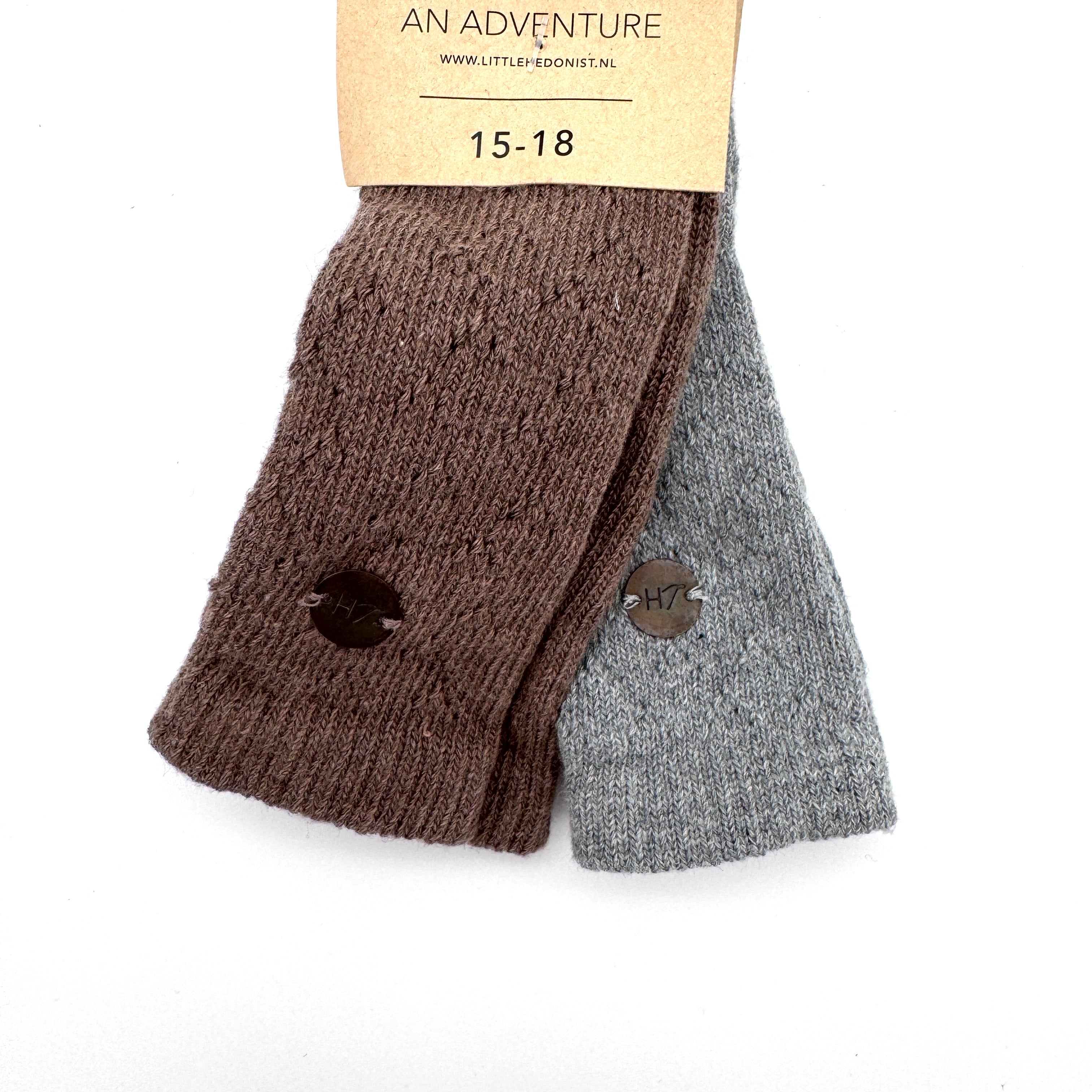 Zwei Paar gerippte Socken, eine braune und eine graue, liegen nebeneinander und tragen oben ein Pappschild mit der Aufschrift AN ADVENTURE und 15-18. Jede Socke hat einen Größenaufkleber mit der Aufschrift H7.