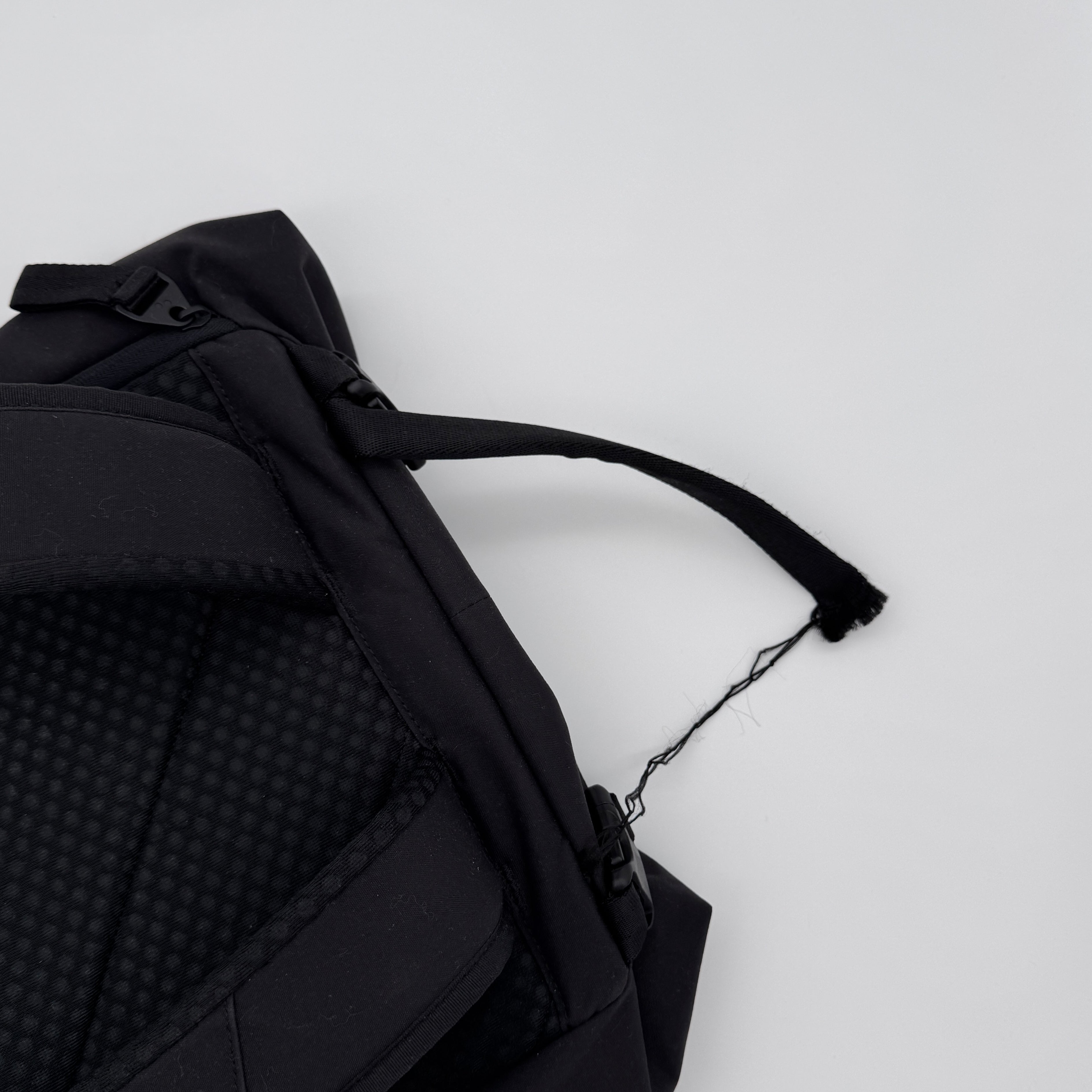Ein schwarzer Rucksack mit einem ausgefransten und teilweise zerrissenen Schultergurt ist vor einem schlichten weißen Hintergrund abgebildet.