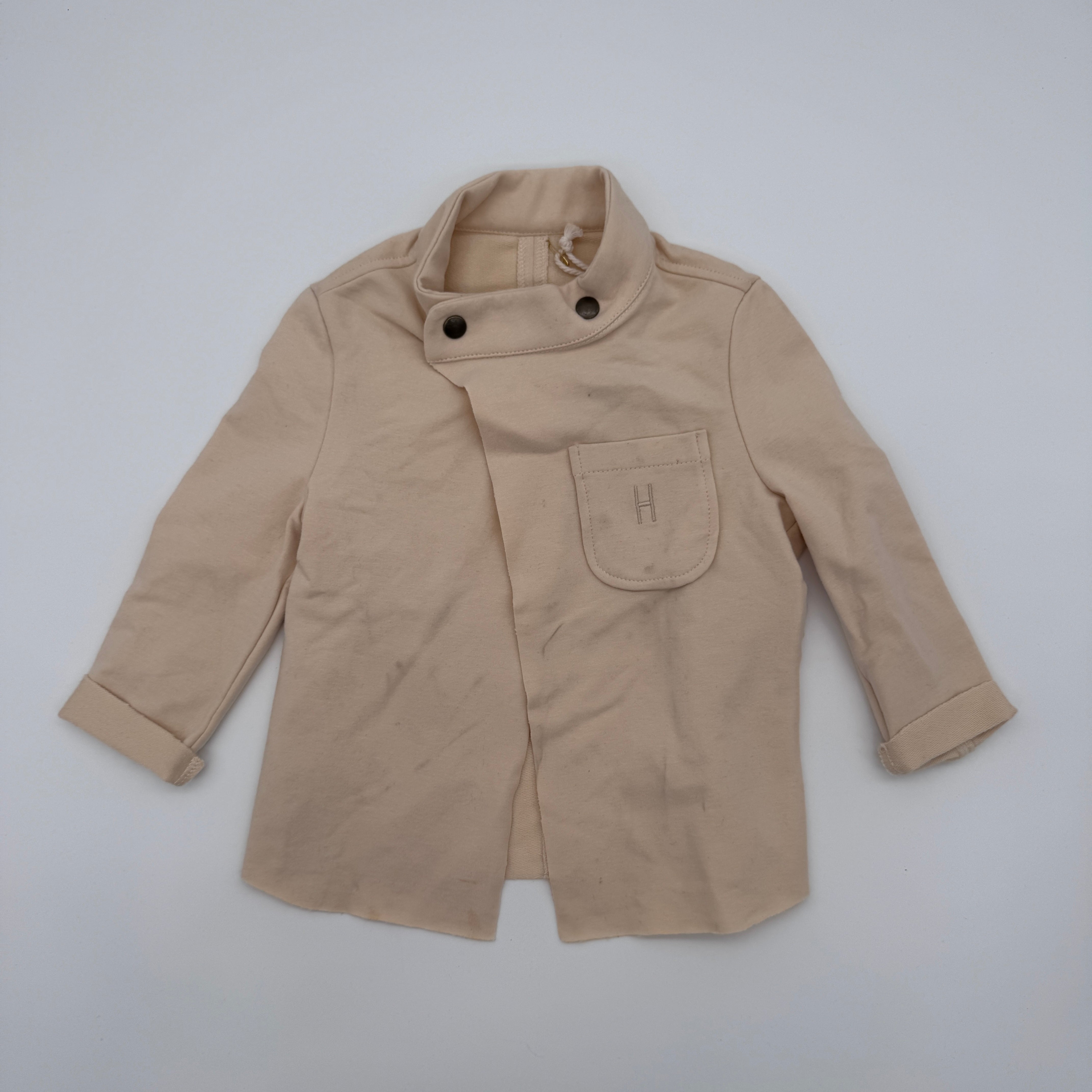 Hellbeige, langärmelige Kinderjacke mit hohem Kragen, zwei schwarzen Knöpfen im Nacken und einer Brusttasche mit aufgesticktem H. Die Ärmel sind leicht aufgekrempelt.