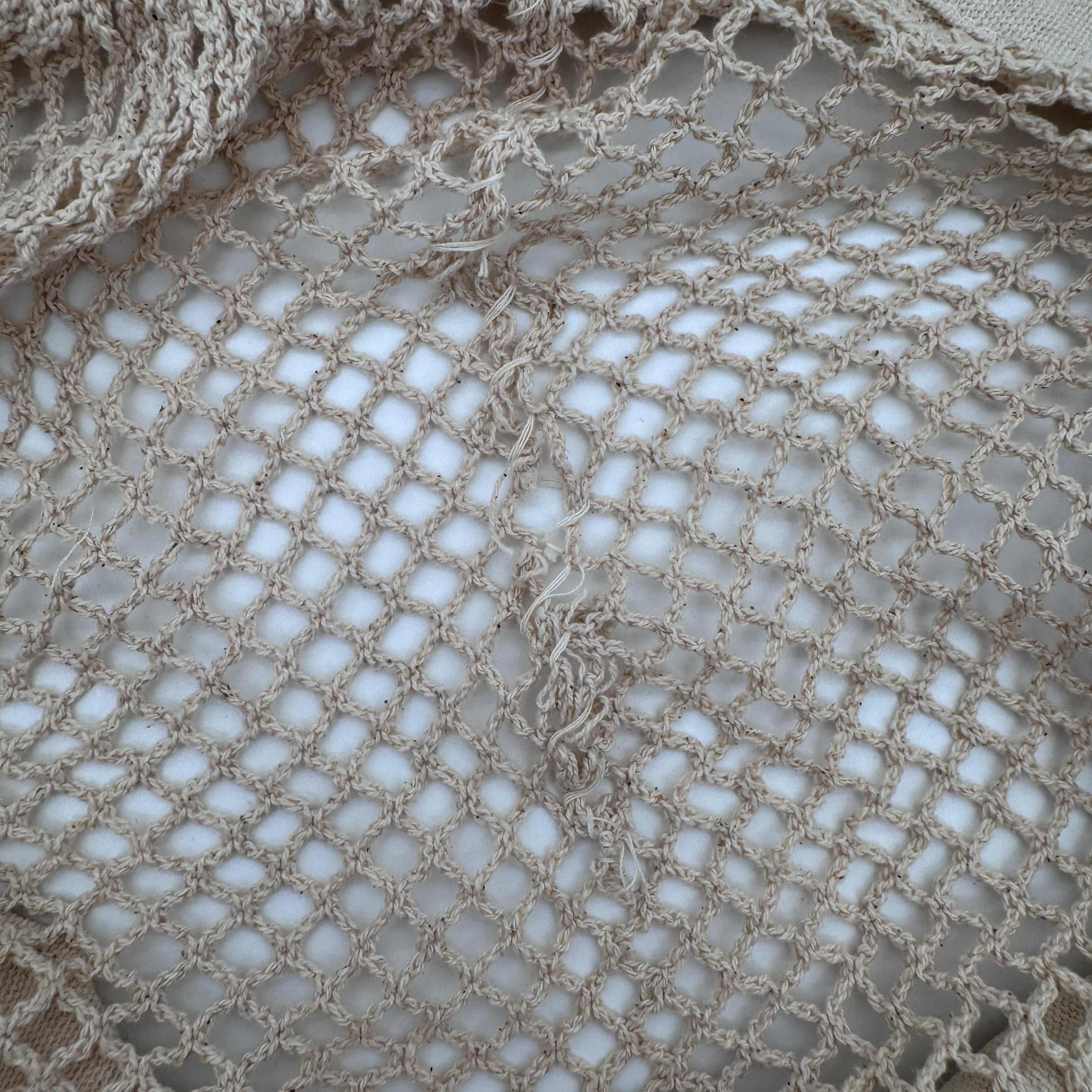 Nahaufnahme eines beigen, locker gewebten Netz- oder Häkelstoffs mit einer sichtbaren Naht oder Unregelmäßigkeit in der Mitte, die das Licht durch das offene, gitterartige Muster hindurchlässt.