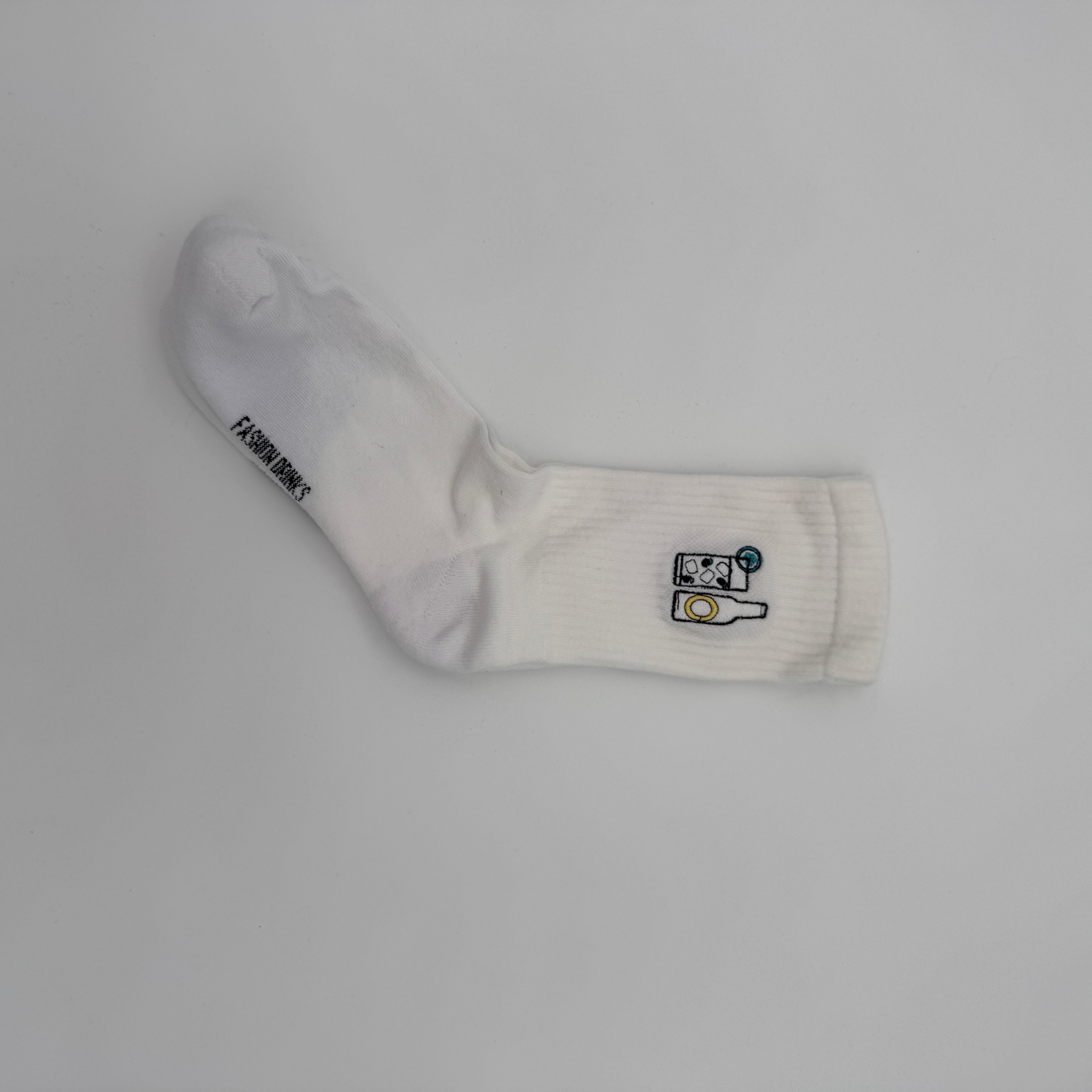 Eine einzelne weiße Socke liegt flach auf einem unifarbenen Hintergrund und ist mit einem kleinen gestickten Motiv einer Kamera und einer gelben Filmrolle in der Nähe des Knöchels versehen. Auf dem Fuß ist das Wort FASHIONSOCKS aufgedruckt.