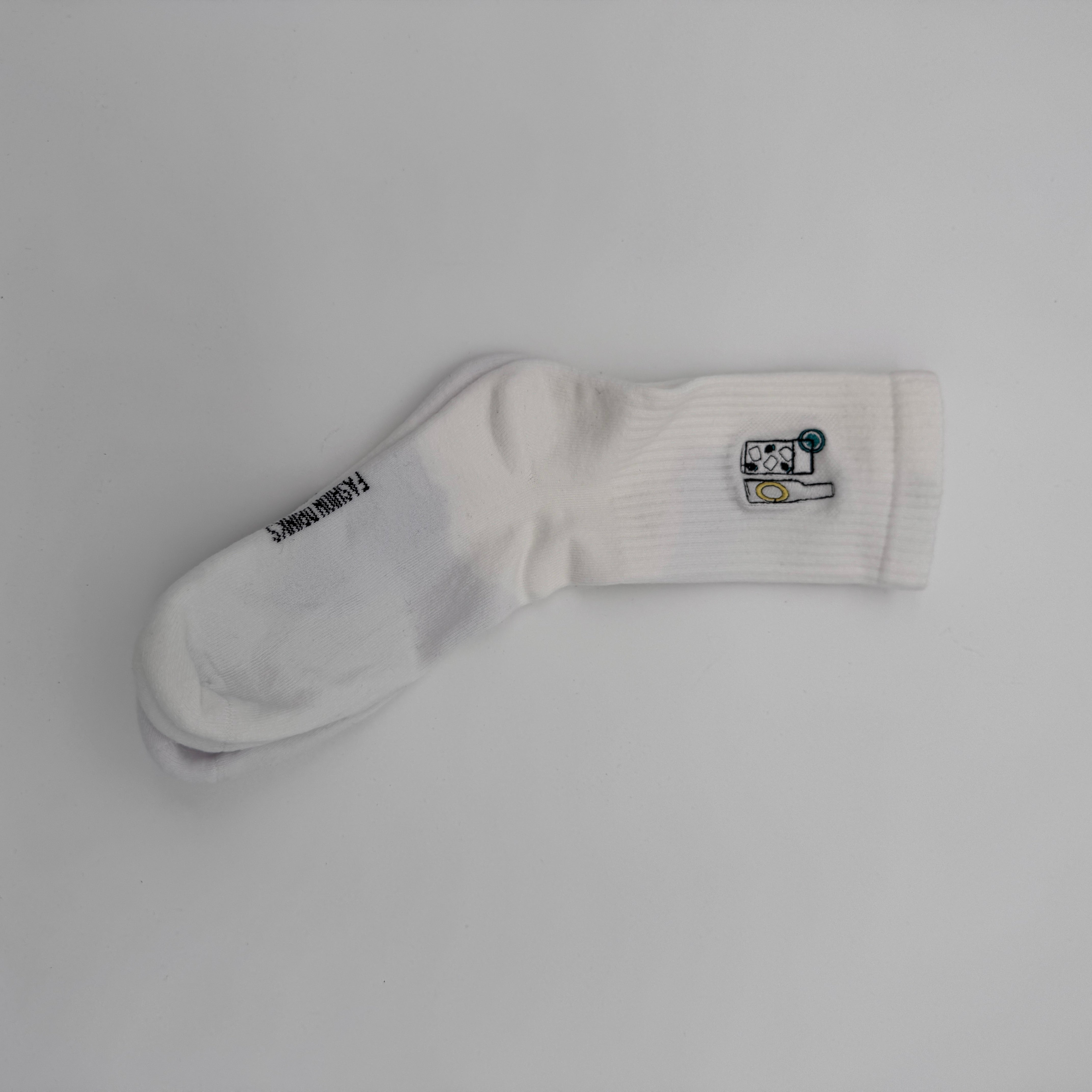 Eine einzelne weiße Socke mit einem kleinen gestickten Waschmaschinen- und gelben Wäschemotiv im oberen Bereich und dem Text FREAKINWASH auf der Sohle auf einem schlichten weißen Hintergrund.