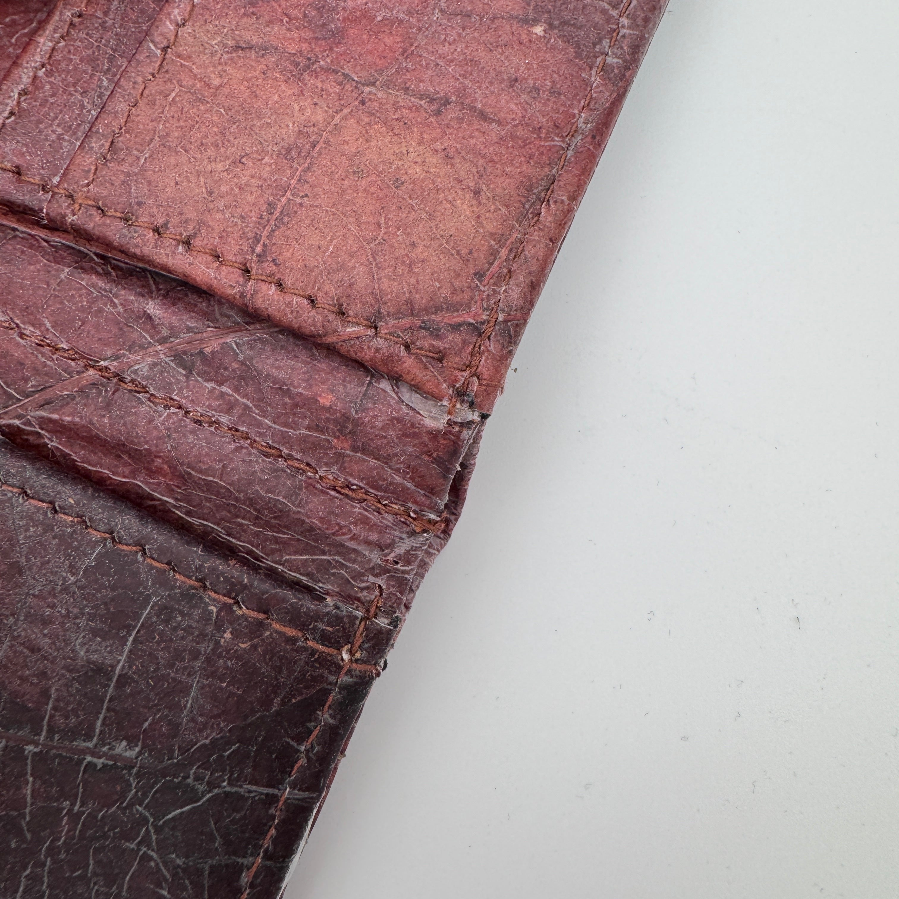 Nahaufnahme einer abgenutzten, rötlich-braunen Lederbörse mit sichtbaren Falten, Nähten und verblassten Stellen auf einer hellgrauen Oberfläche.