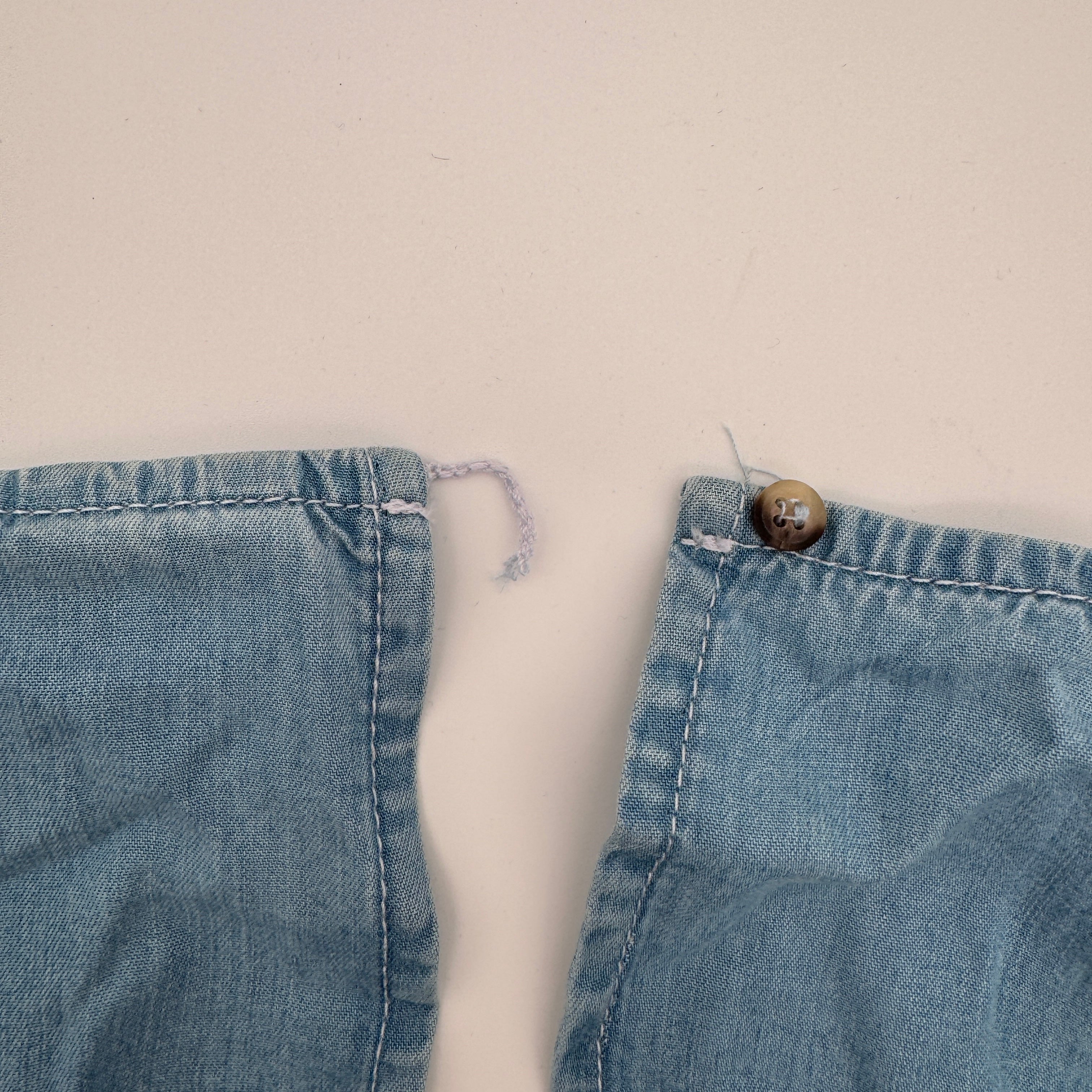Eine Nahaufnahme von zwei Jeanshemd-Manschetten auf einer hellen Fläche. An einer Manschette ist ein loser Faden an der Stelle eines Knopfes zu sehen, an der anderen Manschette ist ein Knopf angenäht.