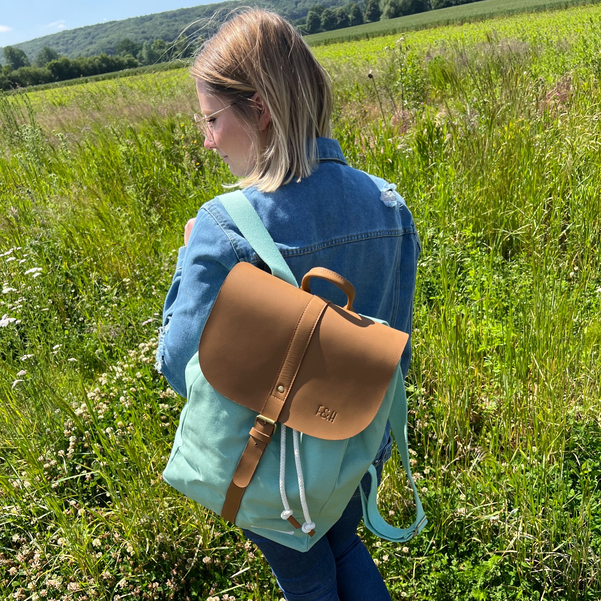 Frau mit Rucksack spaziert durch ein grünes Feld mit Hügeln im Hintergrund, bekleidet mit einer Jeansjacke.