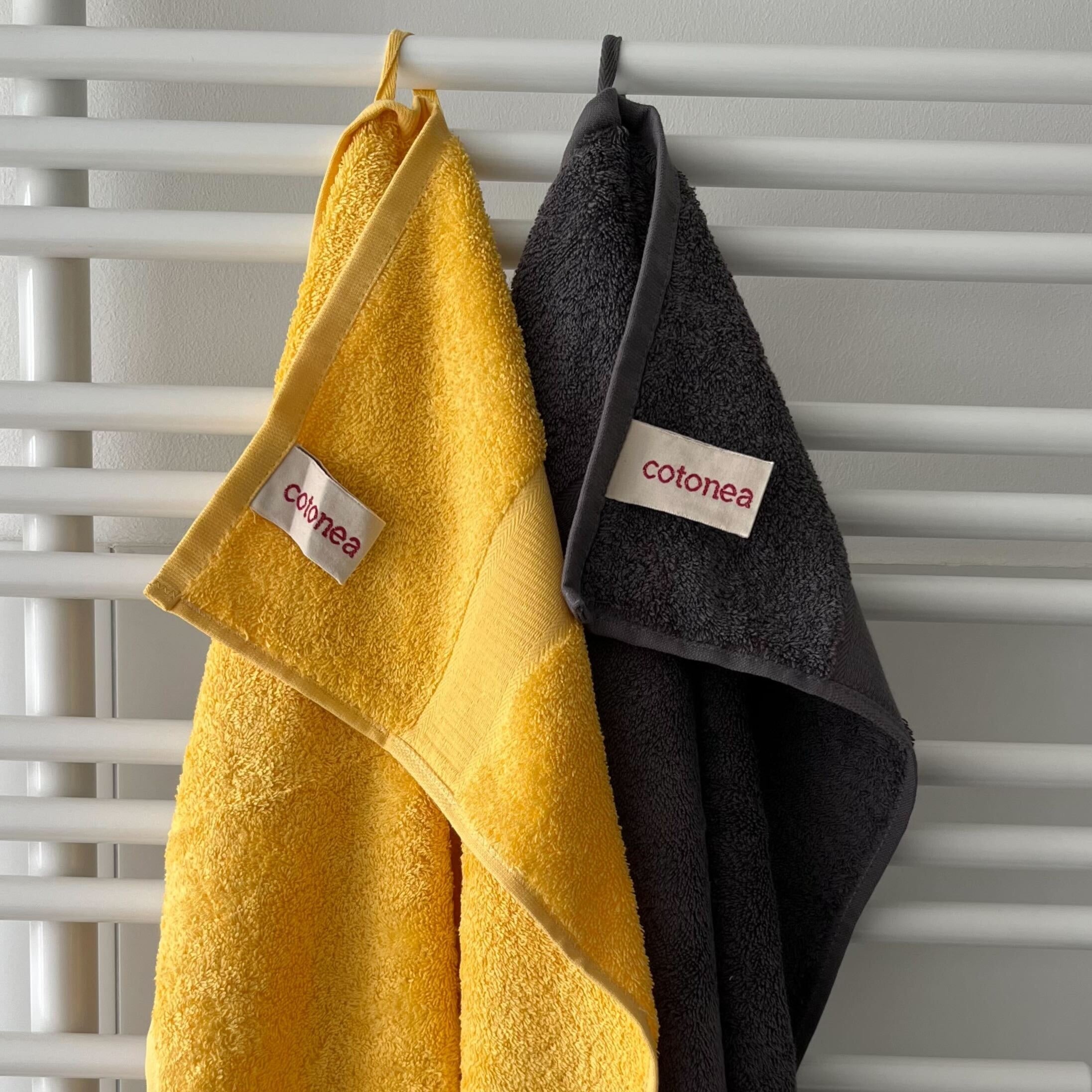 Zwei Handtücher, eines gelb und eines dunkelgrau, hängen an einem weißen Handtuchhalter, beide mit "cotonea"-Etiketten.