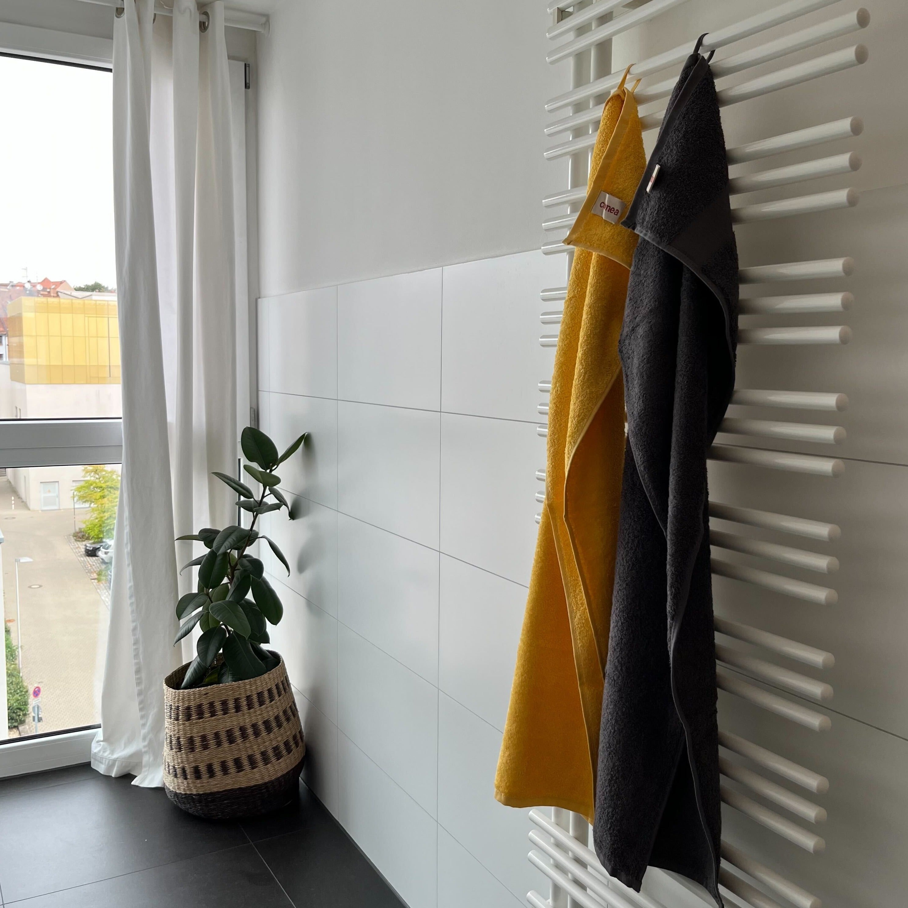 Zwei Handtücher hängen an einem weißen Handtuchwärmer, neben einem Fenster mit Vorhang und einem Korb mit Pflanze.