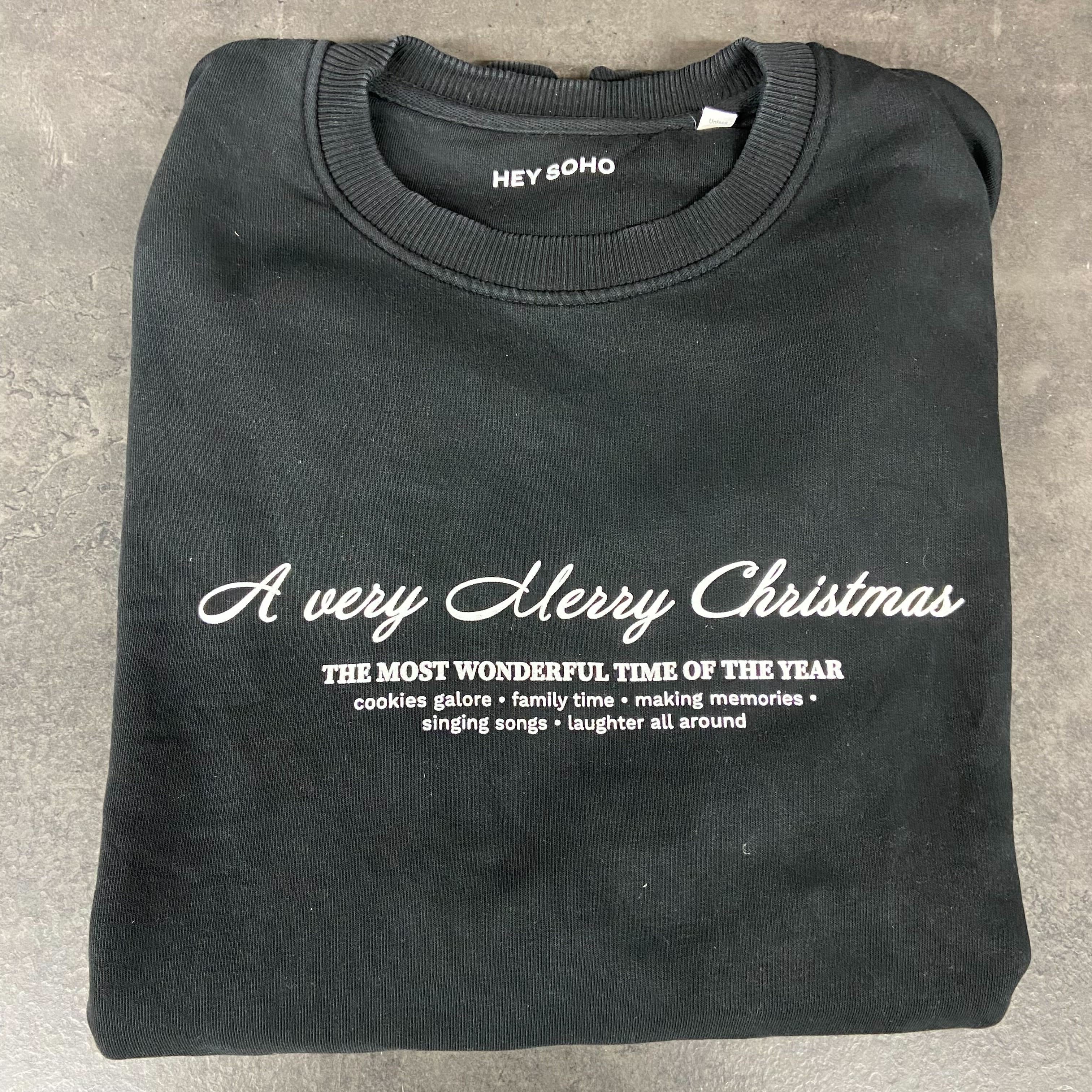 Schwarzer Pullover mit der Aufschrift "A very Merry Christmas" zusammengelegt auf einer grauen Oberfläche.