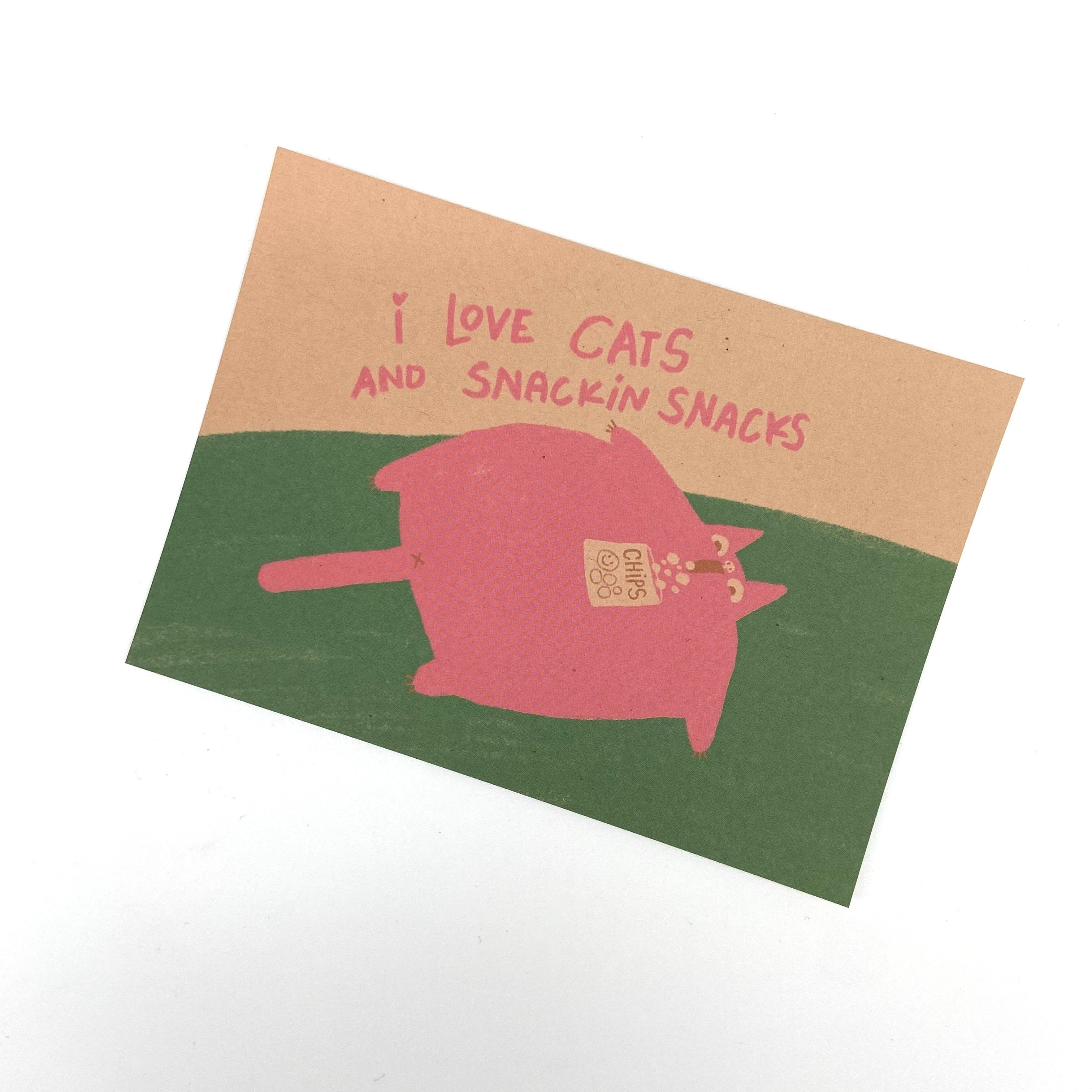 Eine Postkarte zeigt eine dicke rosa Katze mit einem Chipspaket, umgeben von dem Text "I love cats and snackin snacks".