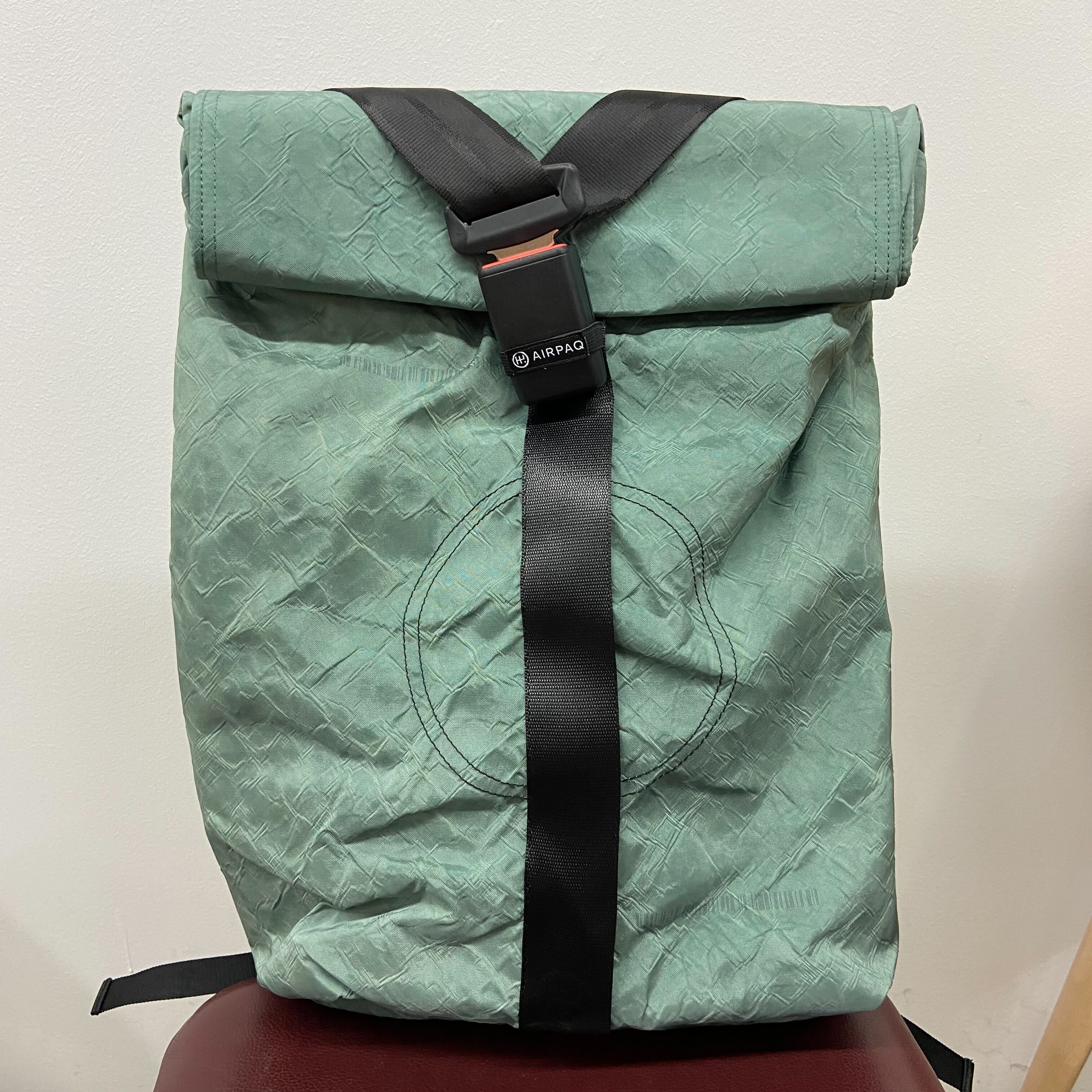 Grüner Rolltop-Rucksack mit schwarzem Gurtband und Schnalle, auf einem roten Stuhl vor einer weißen Wand.