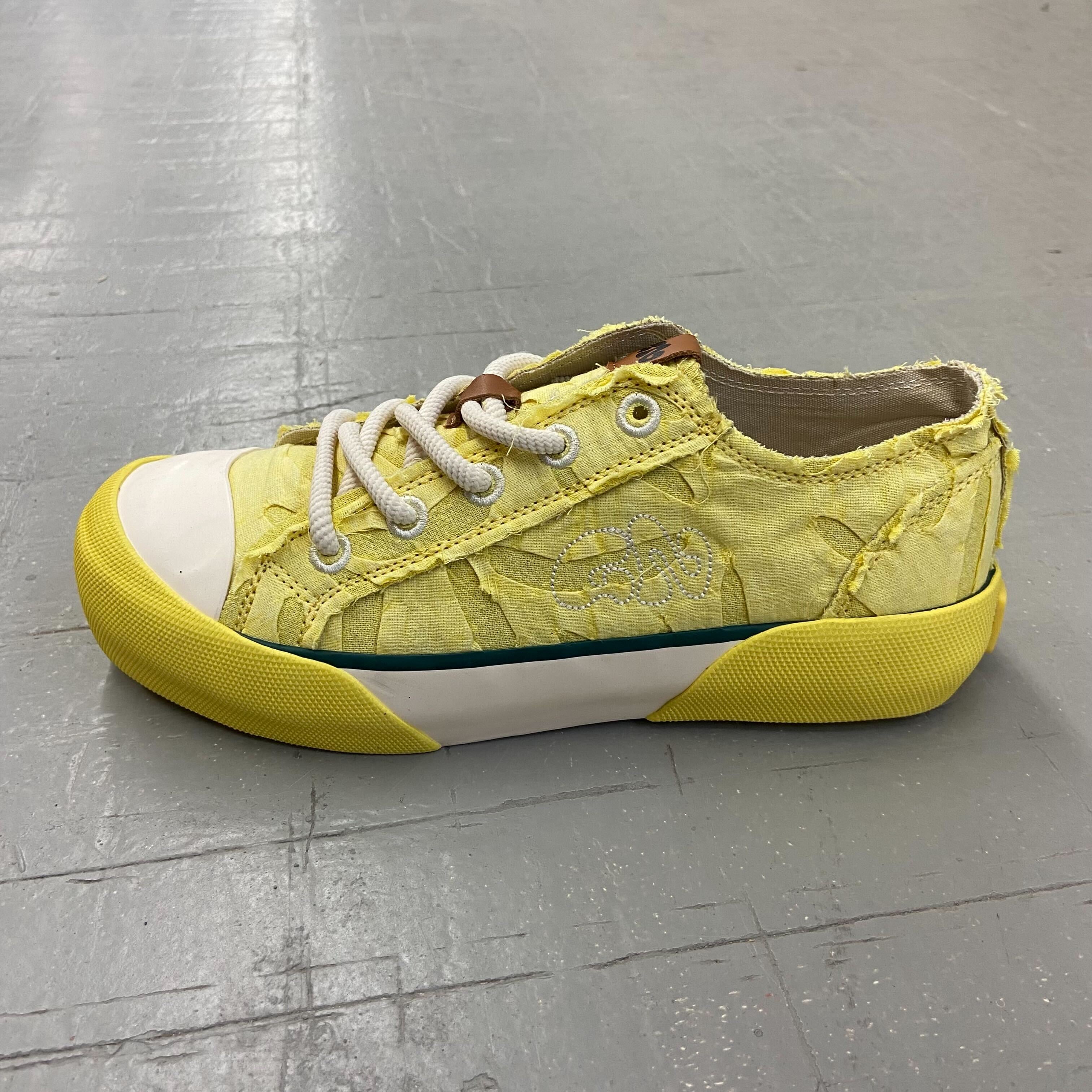 Gelber Sneaker mit weißen Schnürsenkeln und abgenutztem Aussehen auf grauem Boden.