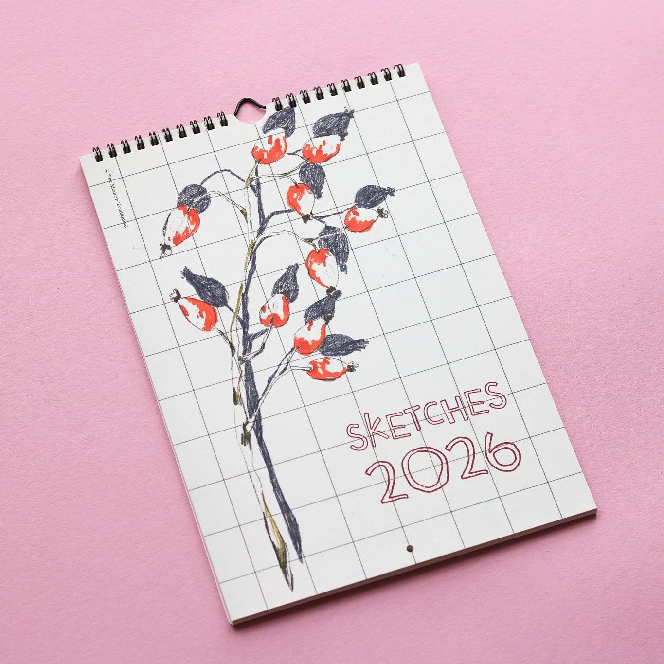 Ein spiralgebundener Kalender mit dem Titel SKETCHES 2026 zeigt eine handgezeichnete botanische Illustration mit roten und schwarzen Beeren auf einem gitterartigen Hintergrund, der sich von einer rosa Oberfläche abhebt.