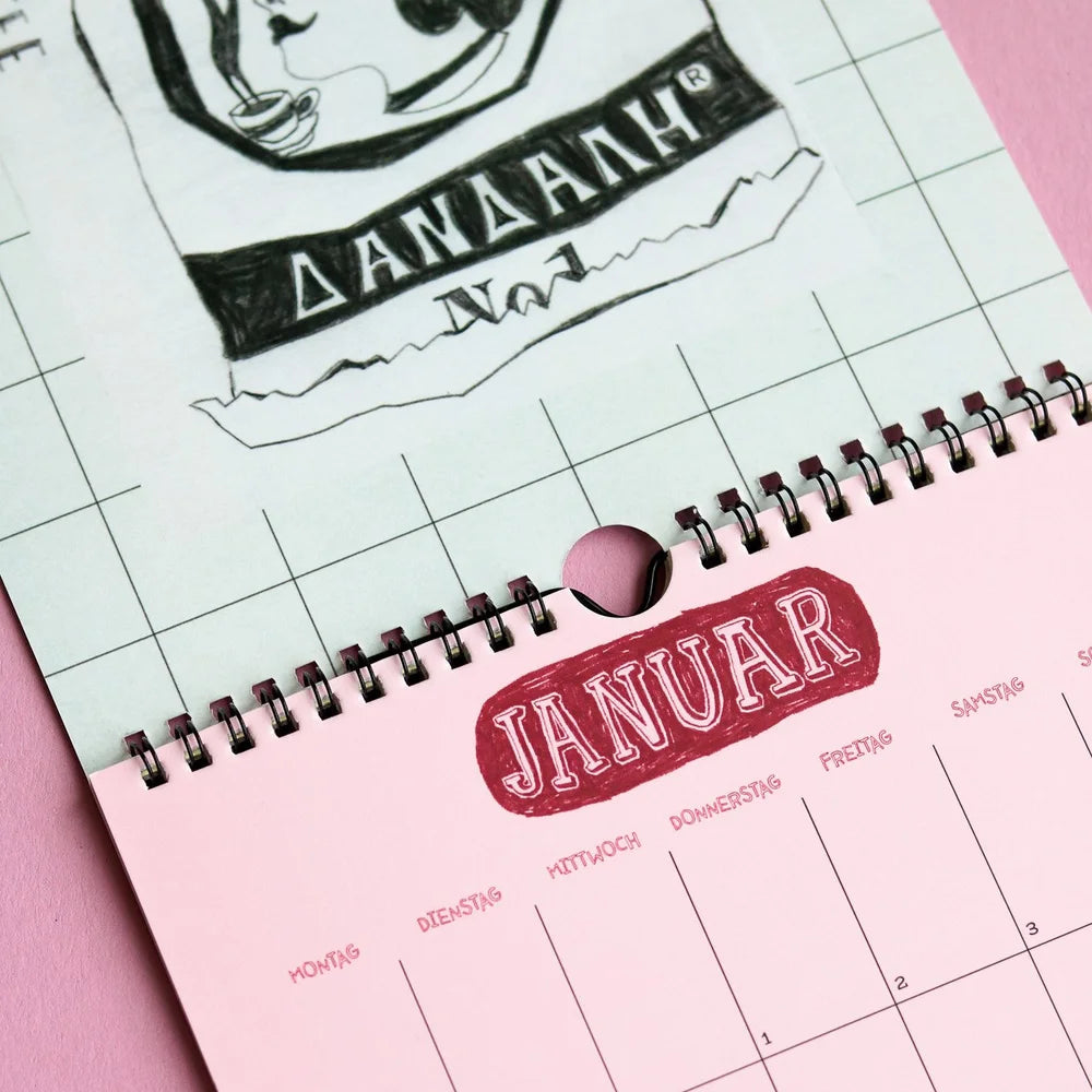 Nahaufnahme eines spiralgebundenen Kalenders, aufgeschlagen im Januar, mit JANUAR in fetten roten Buchstaben und den Wochentagen in deutscher Sprache. Der Hintergrund ist rosa und ein Teil einer Skizze ist auf der vorherigen Seite zu sehen.