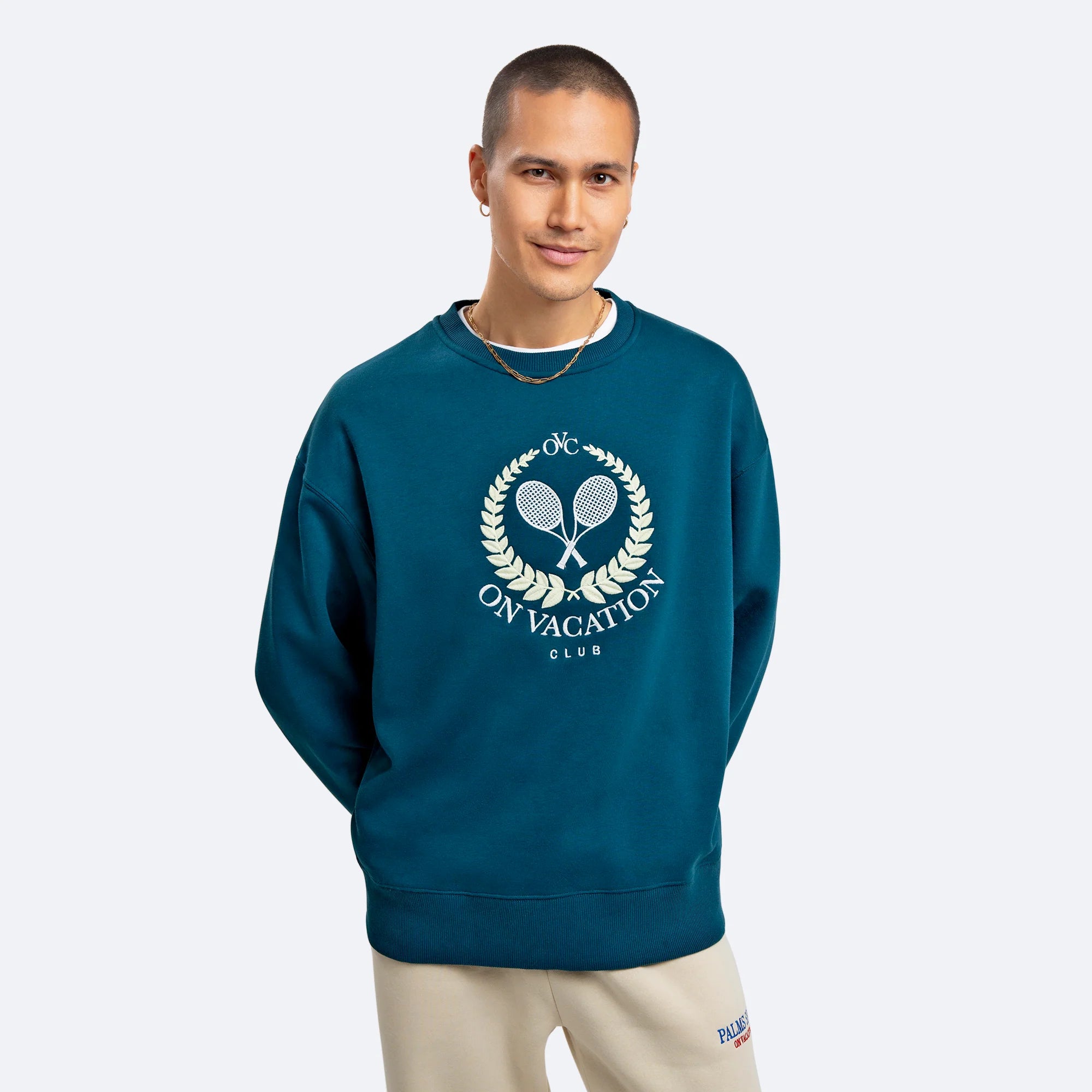 Mann in dunkelblauem Pullover mit Tennisschläger-Logo und Schriftzug "On Vacation Club", lächelt in die Kamera.