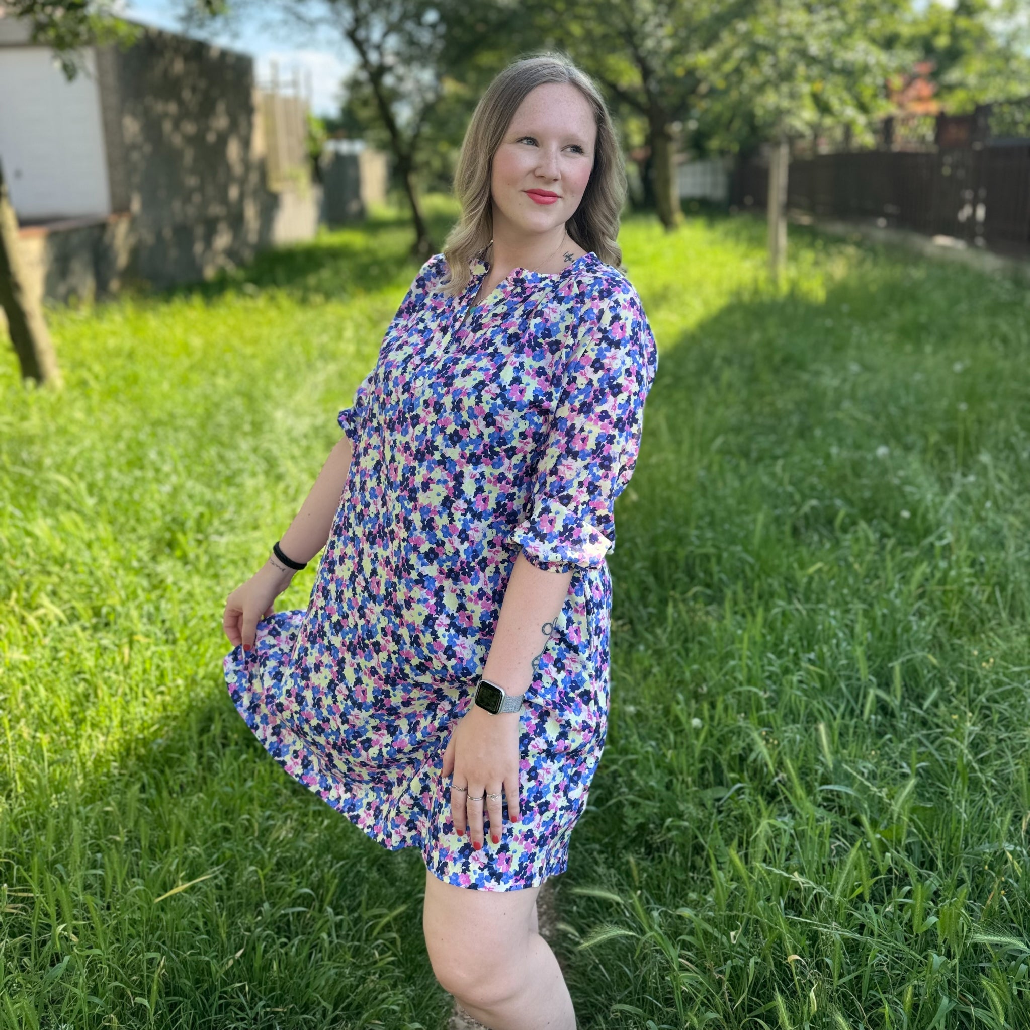 Eine Person in einem bunten Kleid steht auf einer grünen Wiese mit Bäumen im Hintergrund.