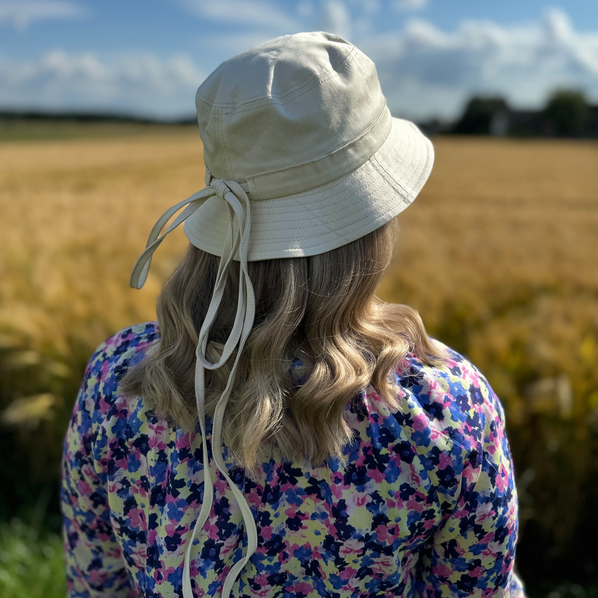 Eine Person mit einem blumigen Oberteil und beigem Hut steht mit dem Rücken zu einem Feld an einem sonnigen Tag.