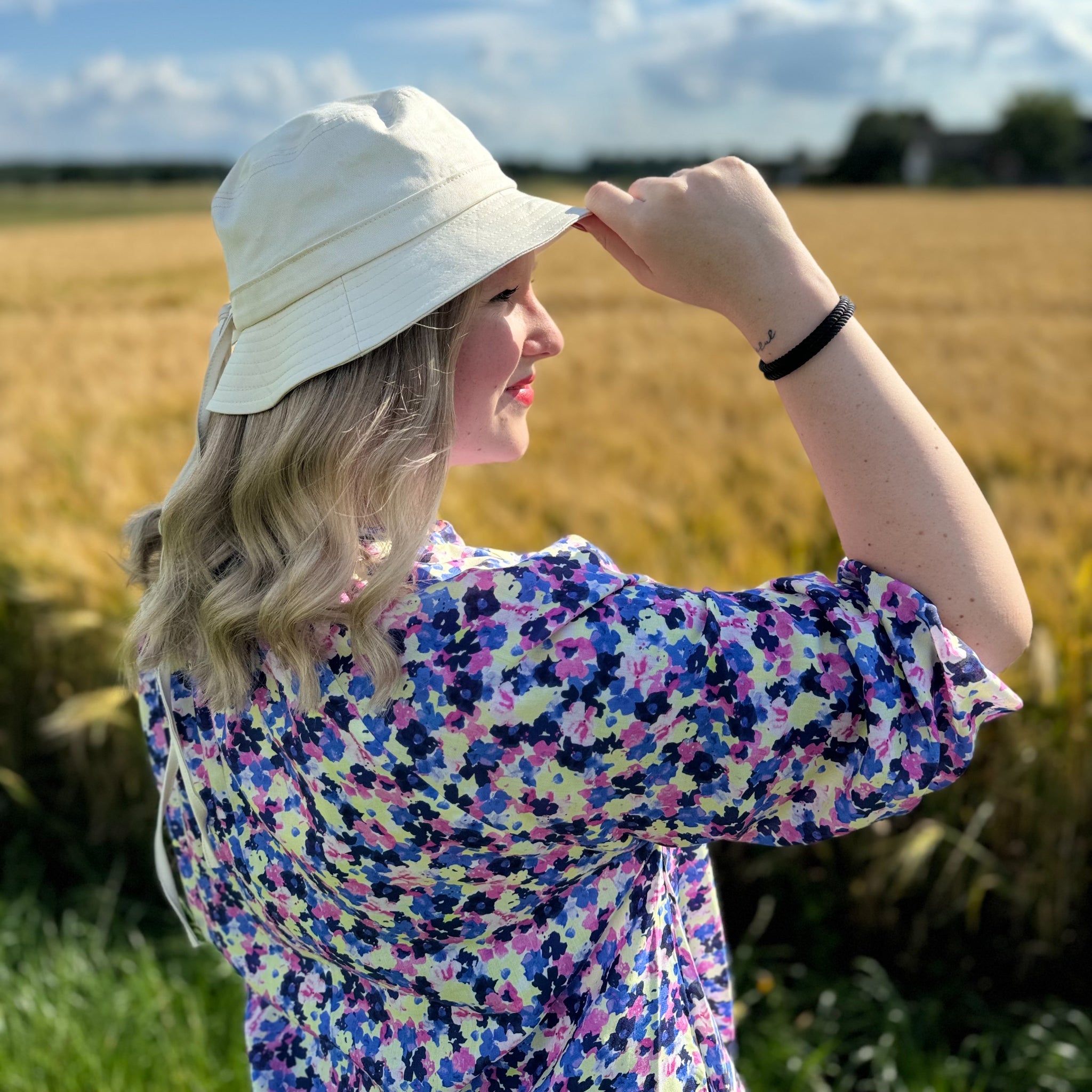 Eine Frau mit einem weißen Hut steht in einem blühenden Feld unter einem blauen Himmel.