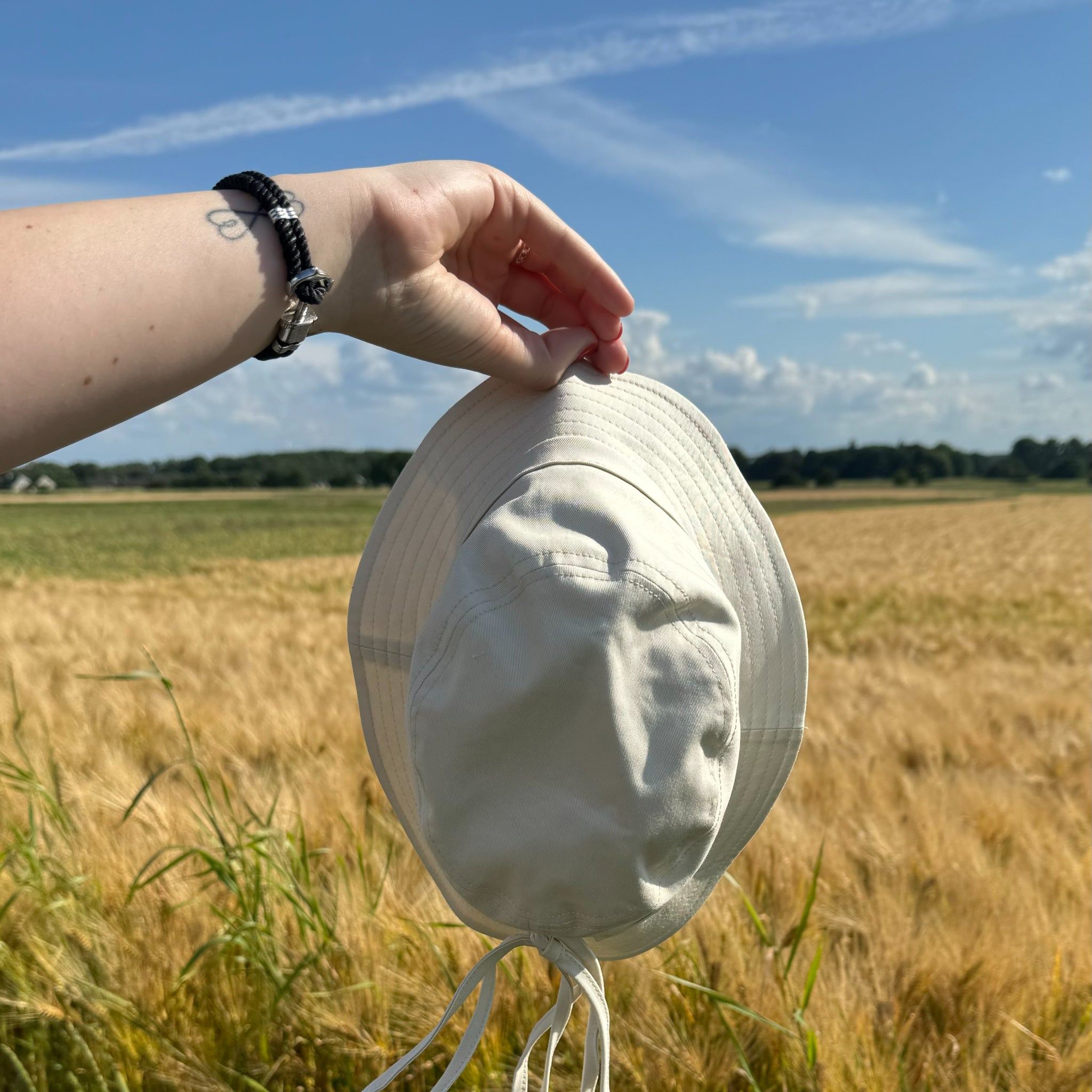 Eine Hand hält einen weißen Hut vor einem goldenen Getreidefeld mit klarem Himmel im Hintergrund.