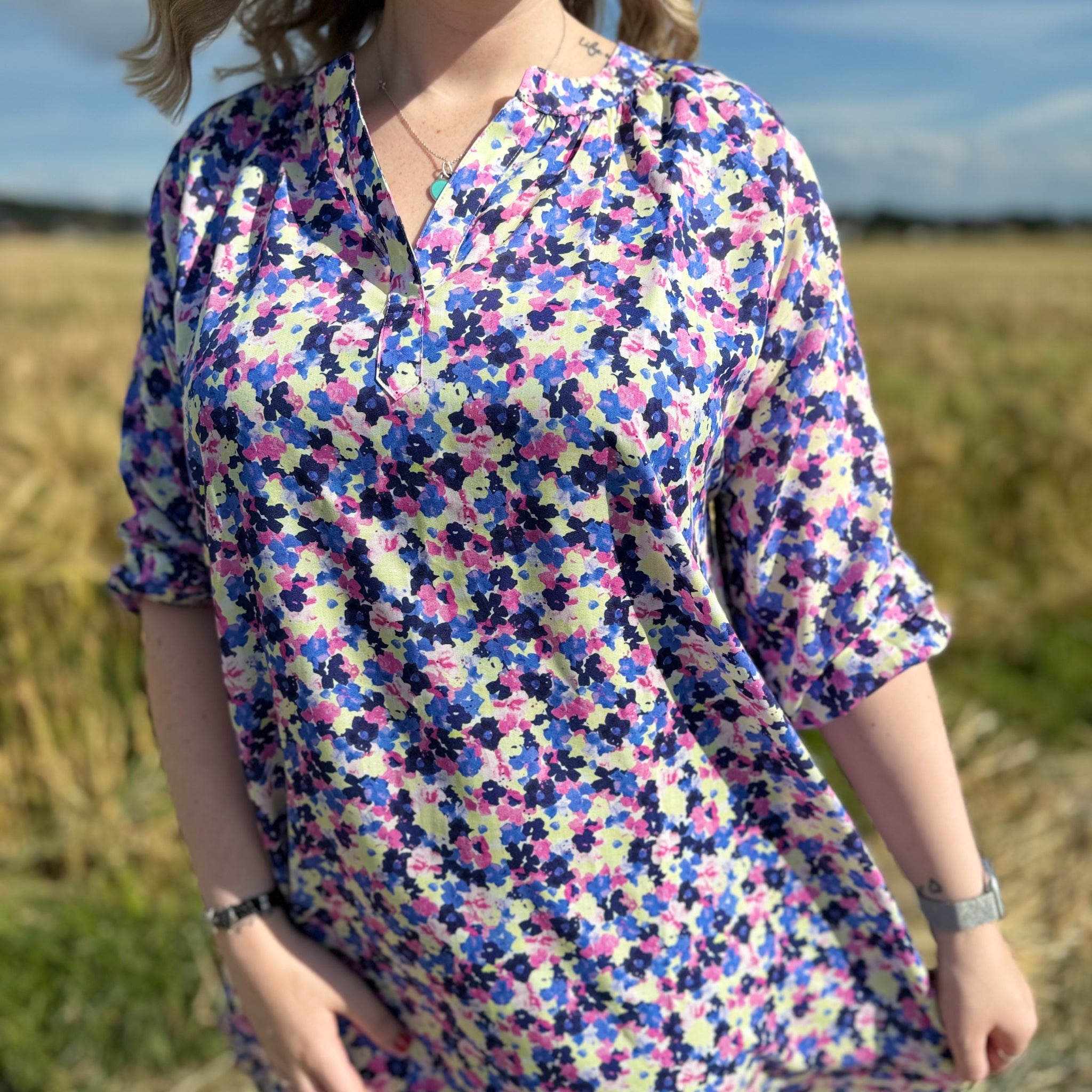 Person in a floral dress stands in a sunlit field, with blue sky in the background.