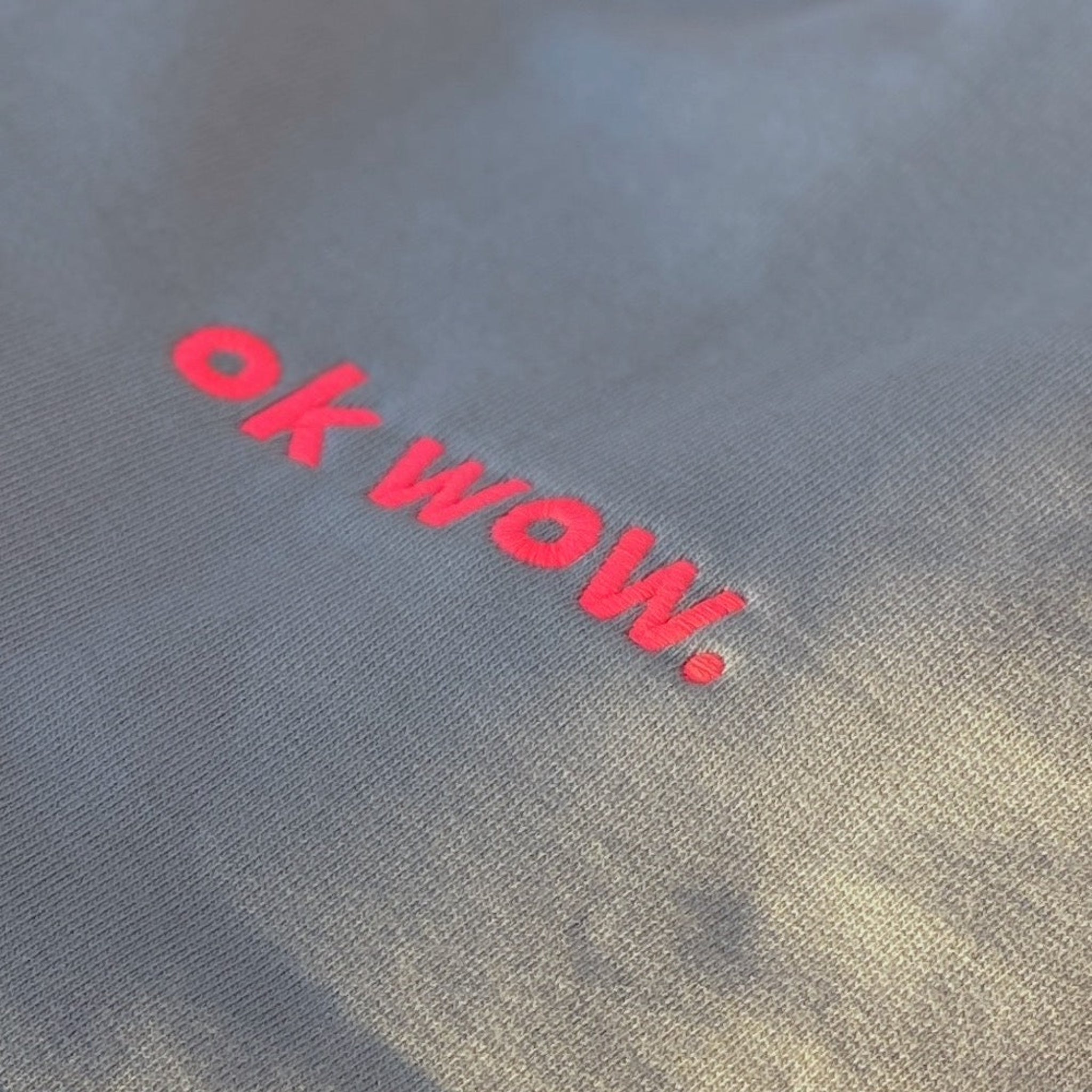 Nahaufnahme eines grauen Stoffes mit den Worten ok wow., die in leuchtend rosa Faden gestickt sind.
