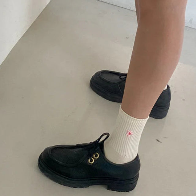 Person mit schwarzen Schuhen, cremefarbenen Socken mit kleinem rosa Muster und einem schwarzen Rock, die auf einem hellen Boden in der Nähe einer weißen Treppe steht.
