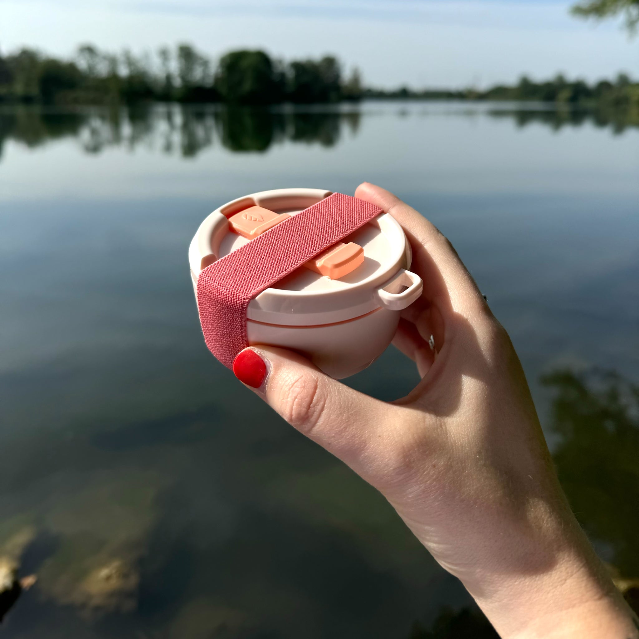 Eine Hand hält eine kleine, runde Box mit rosa Riemen vor einem ruhigen See im Hintergrund.