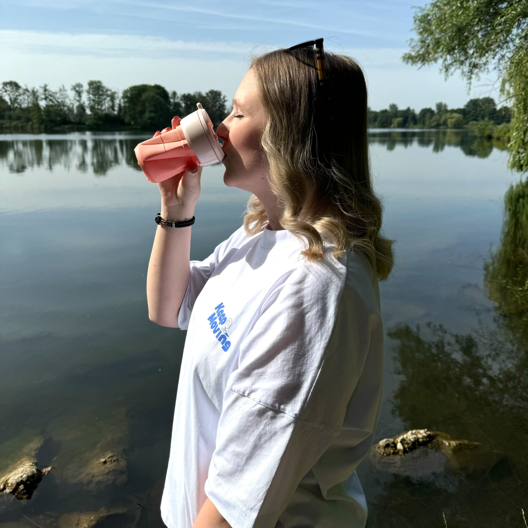 Frau trinkt aus einem Becher am Seeufer, umgeben von ruhigem Wasser und Bäumen.
