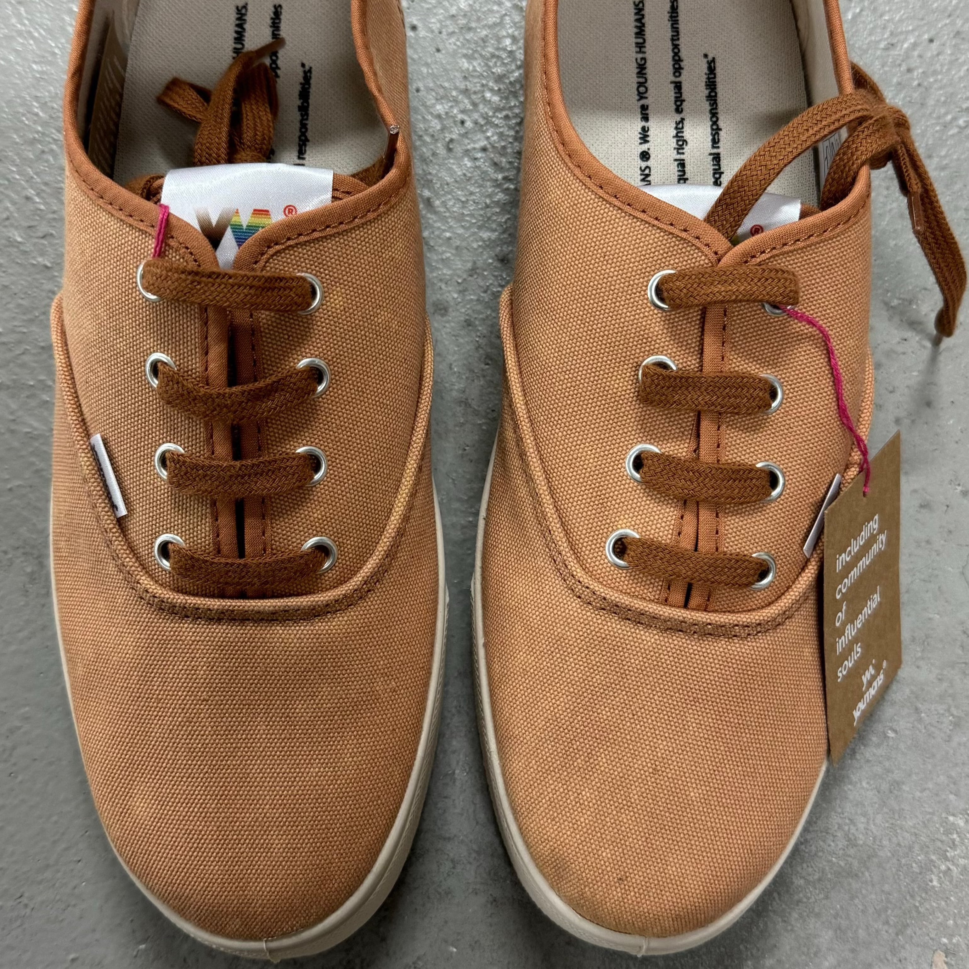 Zwei beige Sneaker mit braunen Schnürsenkeln auf grauem Boden, rechten Schuh mit Etikett am Schnürsenkel.