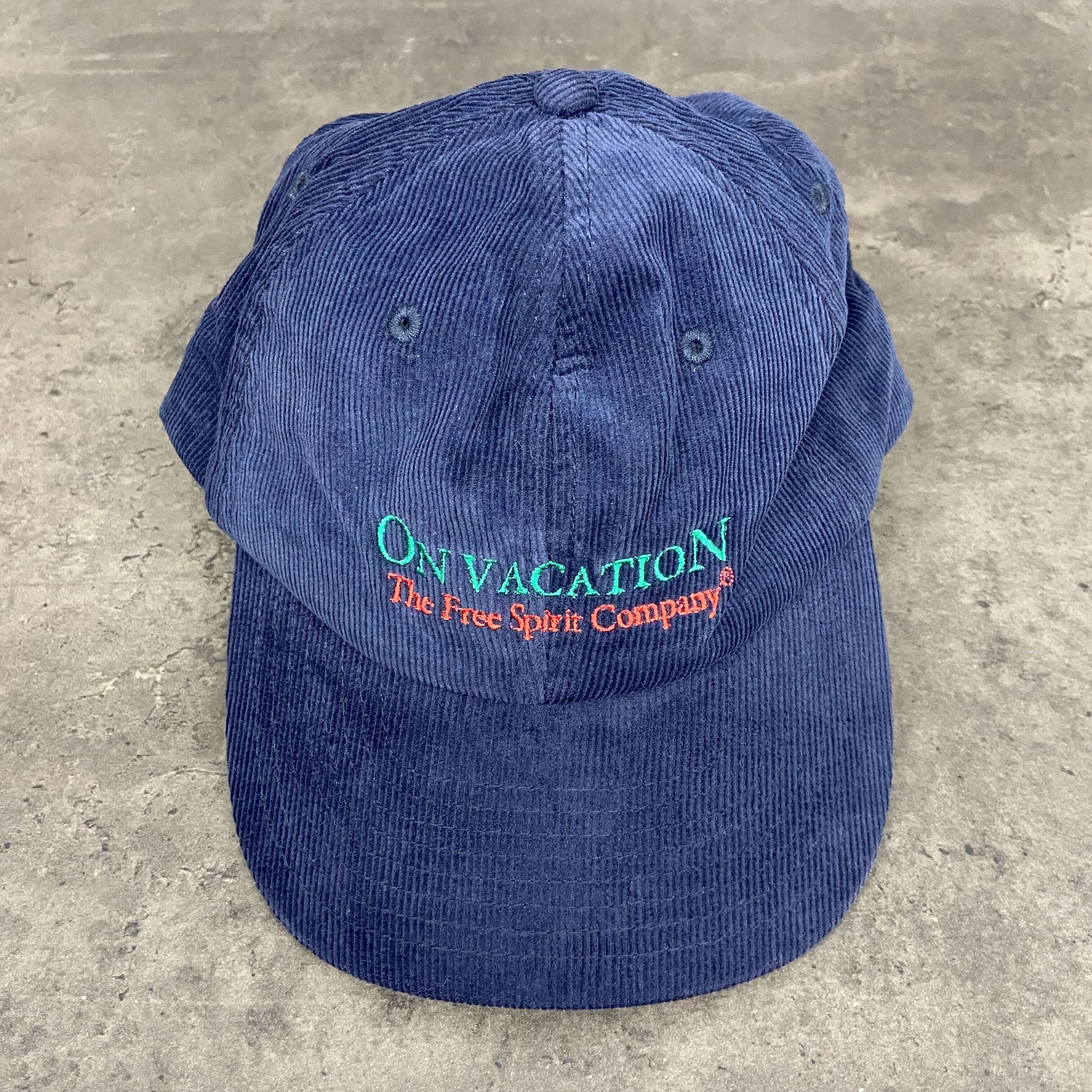 Blaue Kappe mit der Aufschrift "ON VACATION The Free Spirit Company" auf grauem Hintergrund.
