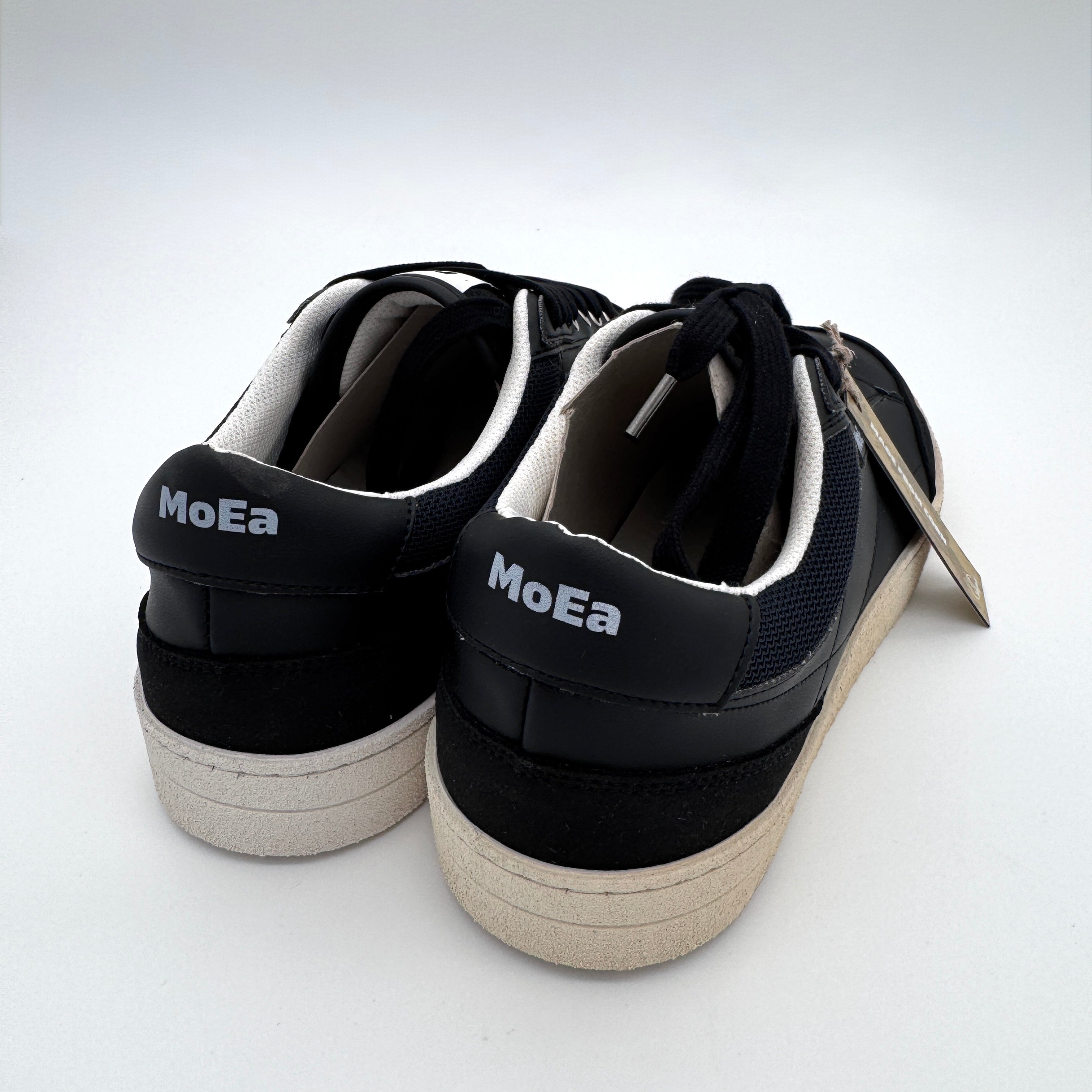 Ein Paar schwarzer Turnschuhe mit weißer Sohle und dem weiß aufgedruckten Markennamen MoEa an der hinteren Ferse, von hinten gesehen vor einem schlichten weißen Hintergrund.
