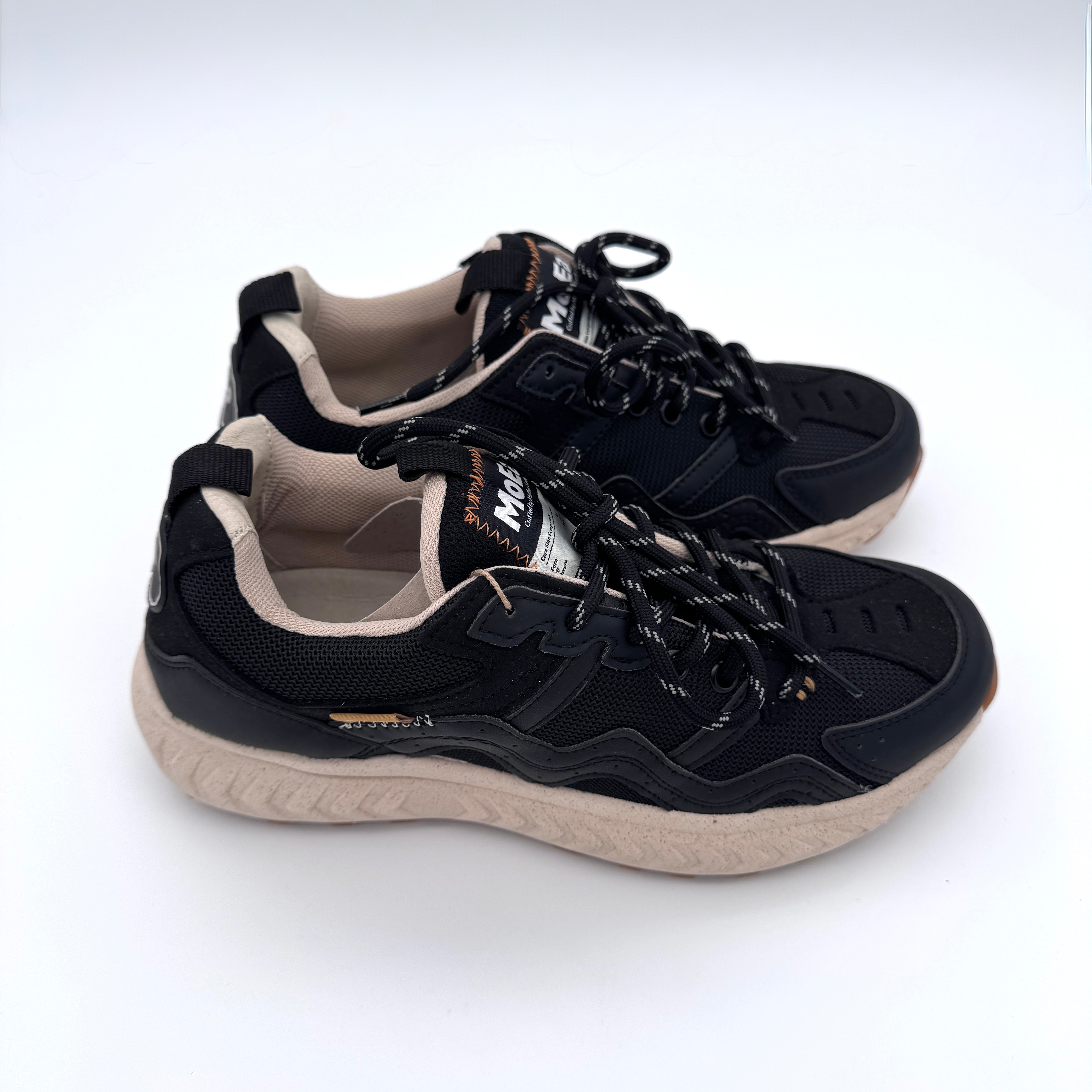 Ein Paar schwarze Turnschuhe mit beiger Sohle und schwarz-weißen Schnürsenkeln, mit beigem Innenfutter und Markenetiketten auf der Zunge, dargestellt auf einem weißen Hintergrund.