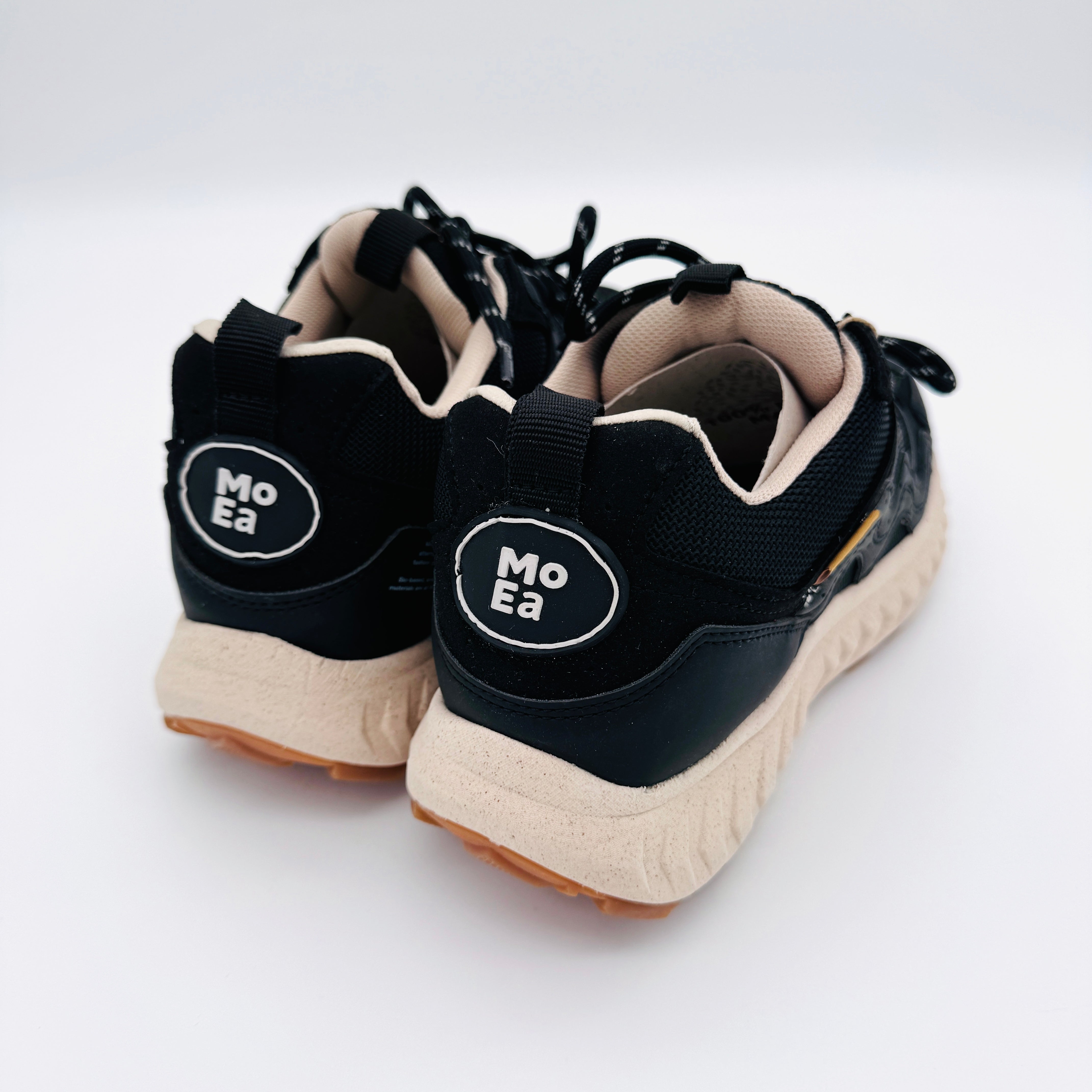 Ein Paar schwarze Turnschuhe mit beiger Sohle, von hinten gesehen, mit ovalen Aufnähern an den Fersen mit dem Text Mo Ea in weißen Buchstaben.