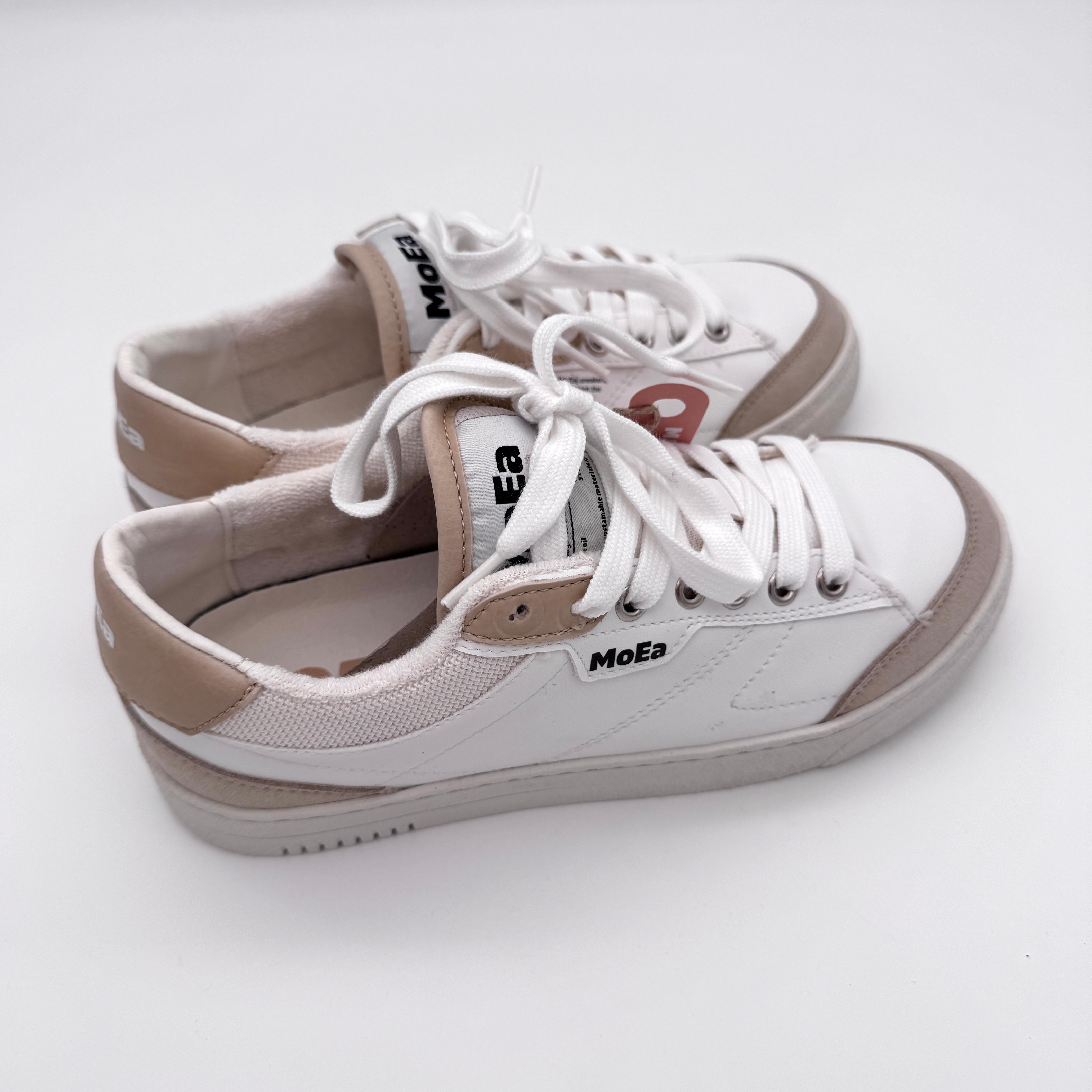 Ein Paar weiße Sneaker mit beigen und hellbraunen Akzenten, weißen Schnürsenkeln und MoEa-Branding an der Seite und auf der Zunge, platziert auf einer weißen Oberfläche.