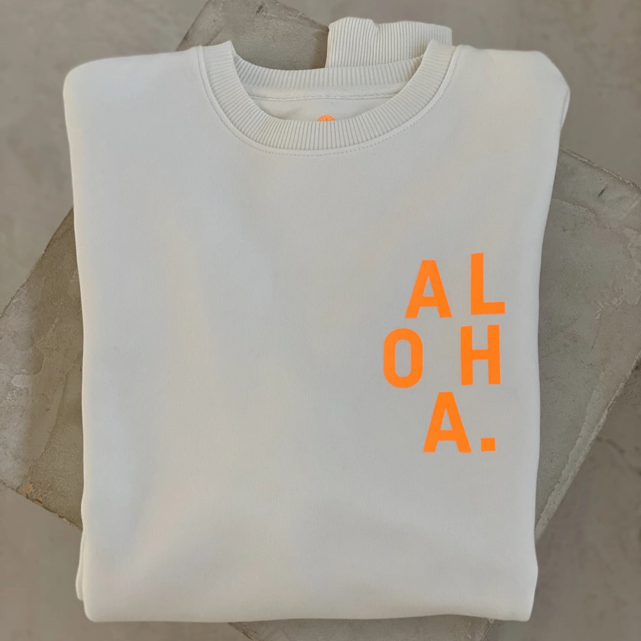 Ein gefaltetes weißes Sweatshirt mit dem Wort "ALOHA." in fetten, leuchtend orangefarbenen Buchstaben auf der Vorderseite, ausgestellt auf einer Betonfläche.
