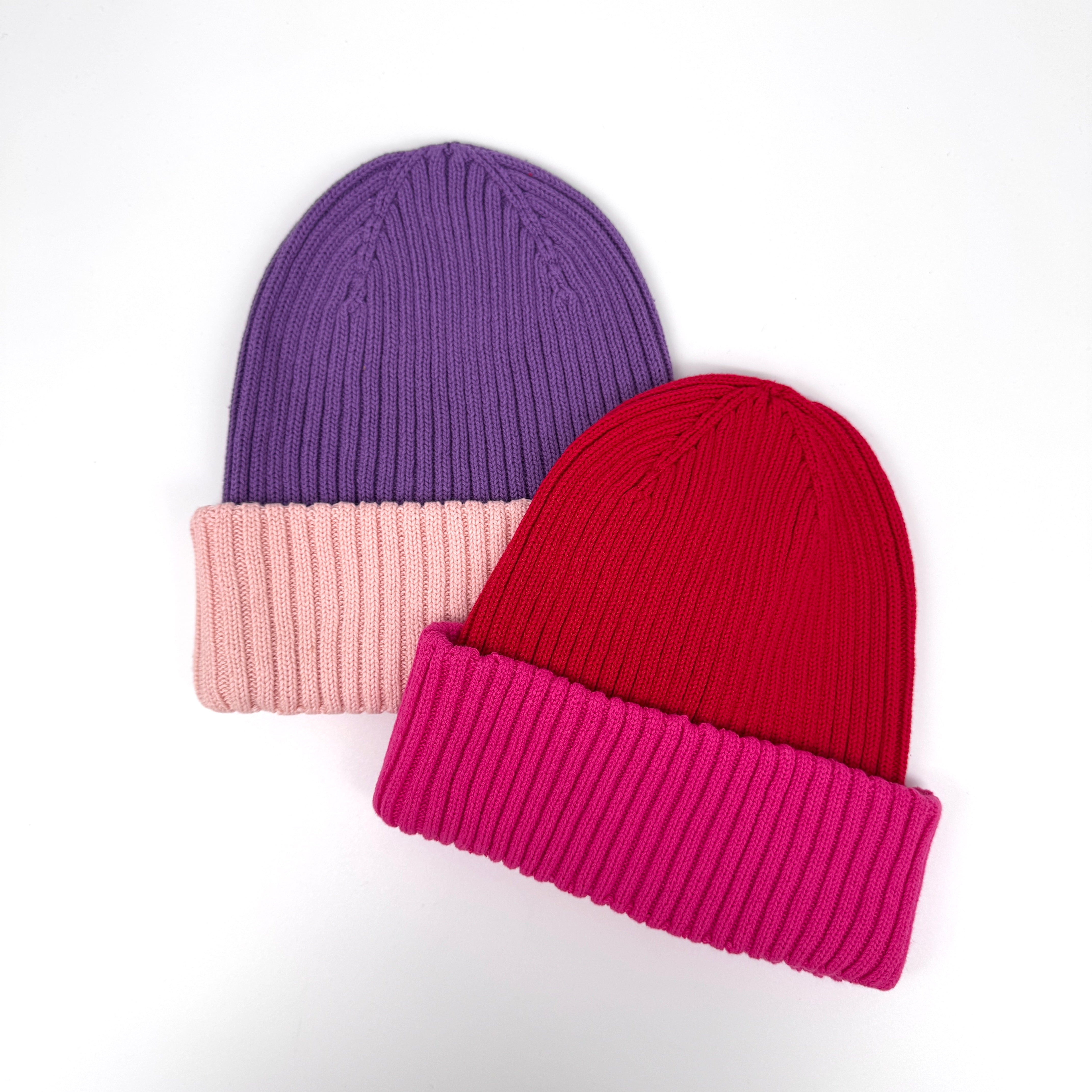 Zwei Rippenstrickmützen auf weißem Grund: eine lila mit hellrosa Bündchen, die andere rot mit hellrosa Bündchen.