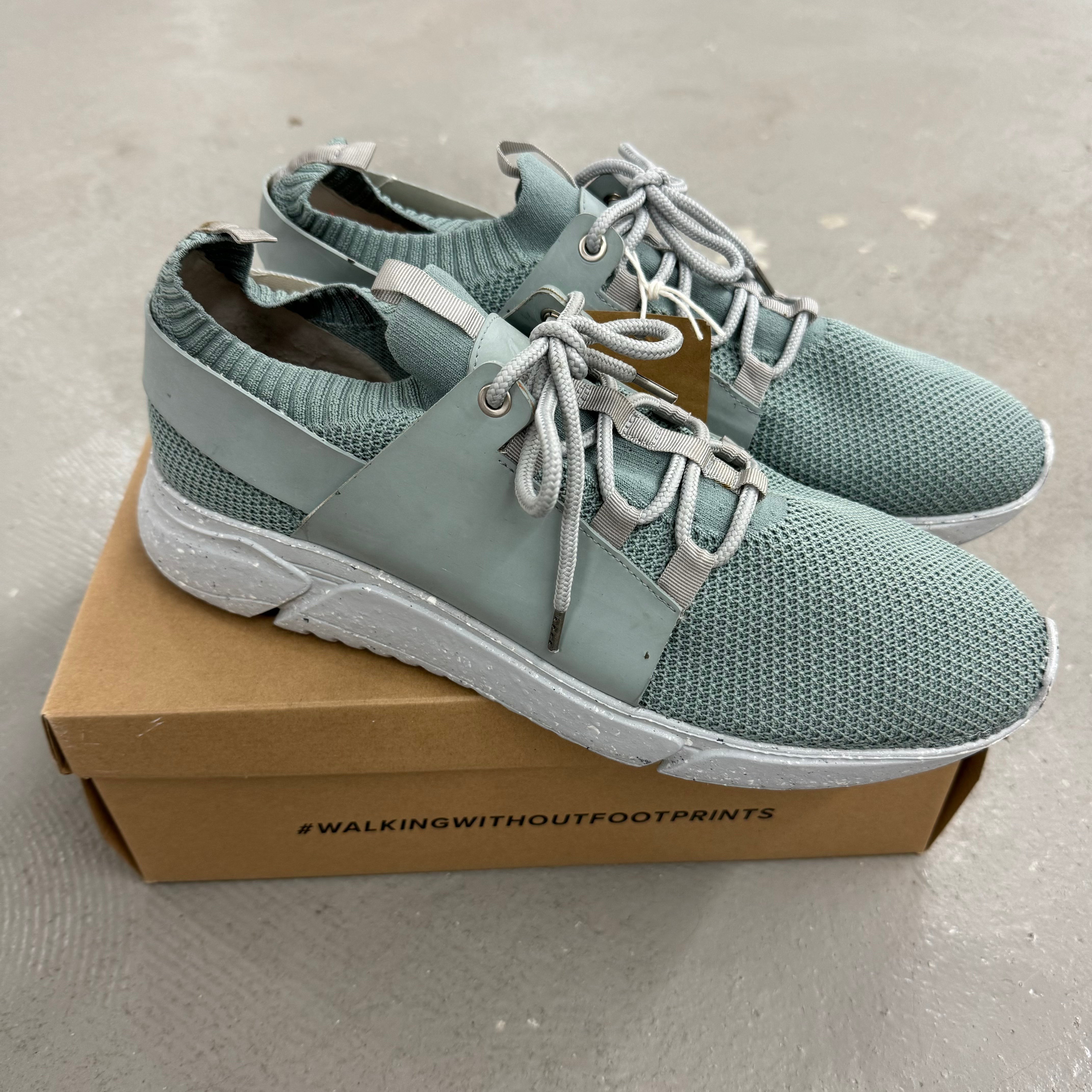 Graue Sportschuhe mit weißer Sohle stehen auf einem braunen Karton mit der Aufschrift "#WALKINGWITHOUTFOOTPRINTS".