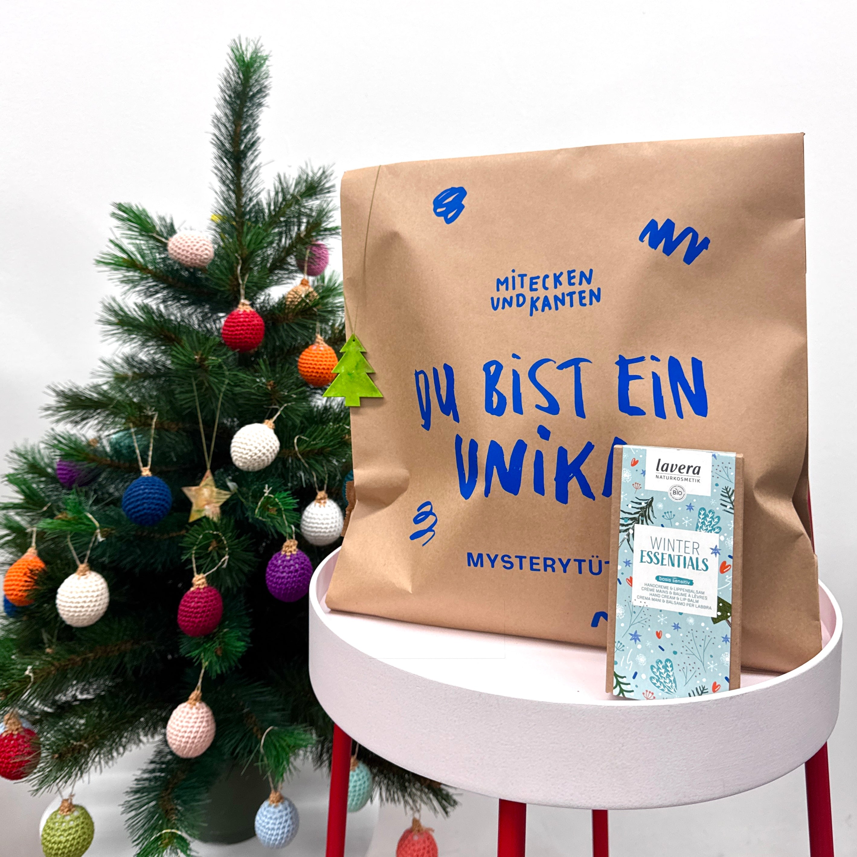 Ein kleiner geschmückter Weihnachtsbaum steht neben einer braunen Papiertüte mit blauer Schrift und einer Schachtel Lavera Winter Essentials auf einem rosa Tisch mit roten Beinen.