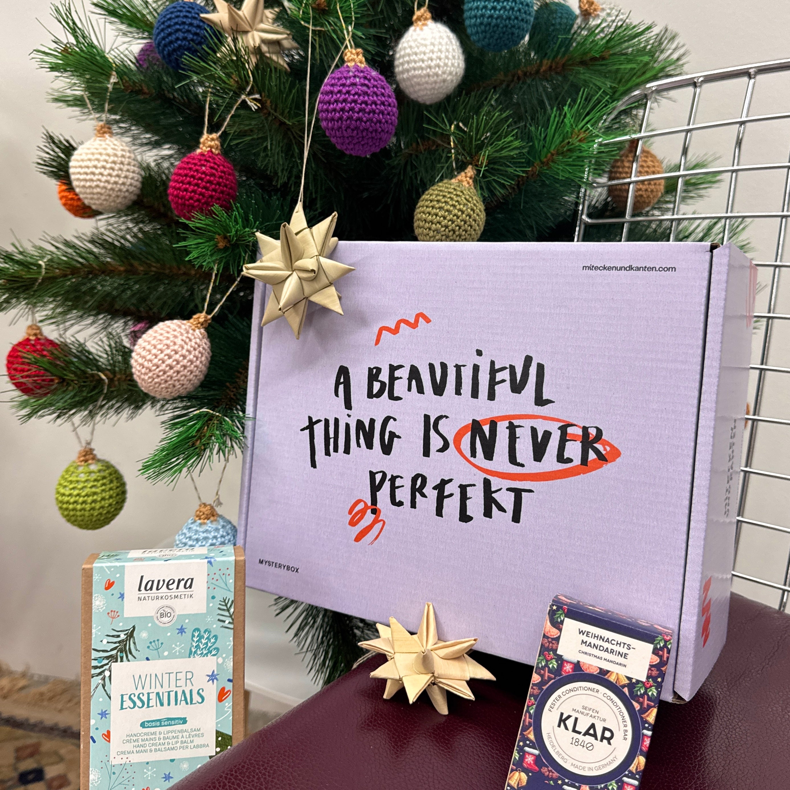 Eine festliche Szene mit einem geschmückten Weihnachtsbaum, einem Drahtstuhl und einer Geschenkbox mit der Aufschrift "A Beautiful Thing Is Never Perfekt". Zwei Geschenkartikel und goldener Sternschmuck sind vor der Schachtel angeordnet.